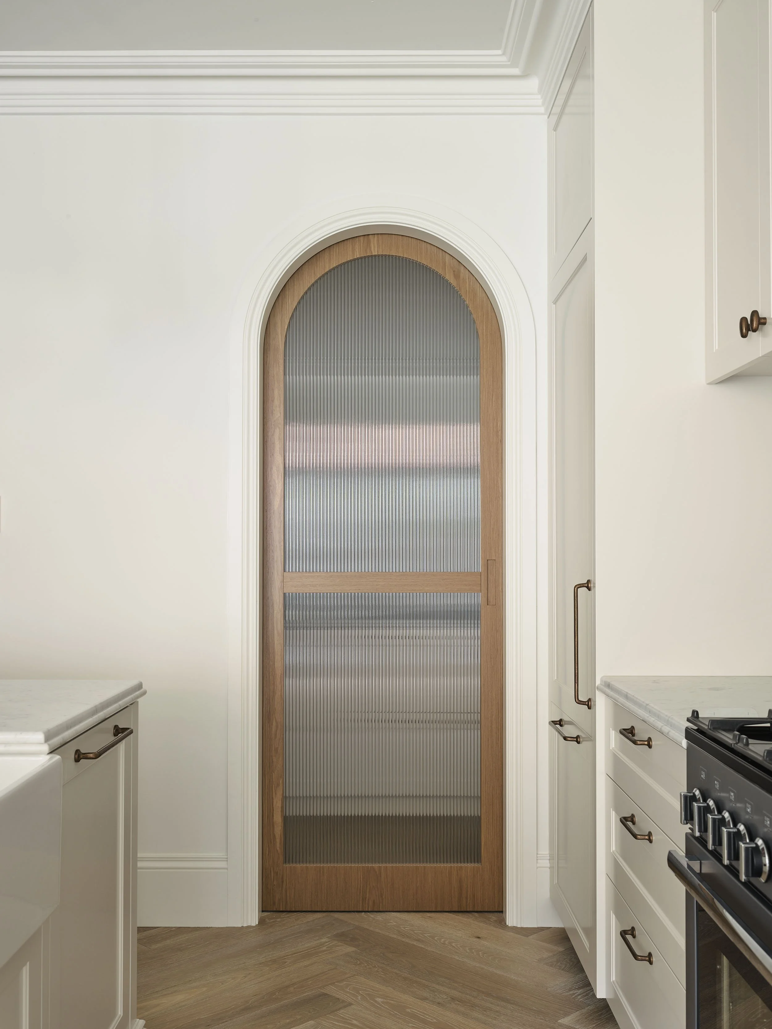 Interior of a kitchen with a wooden and frosted glass arched door, white cabinets, marble countertops, and a gas stove