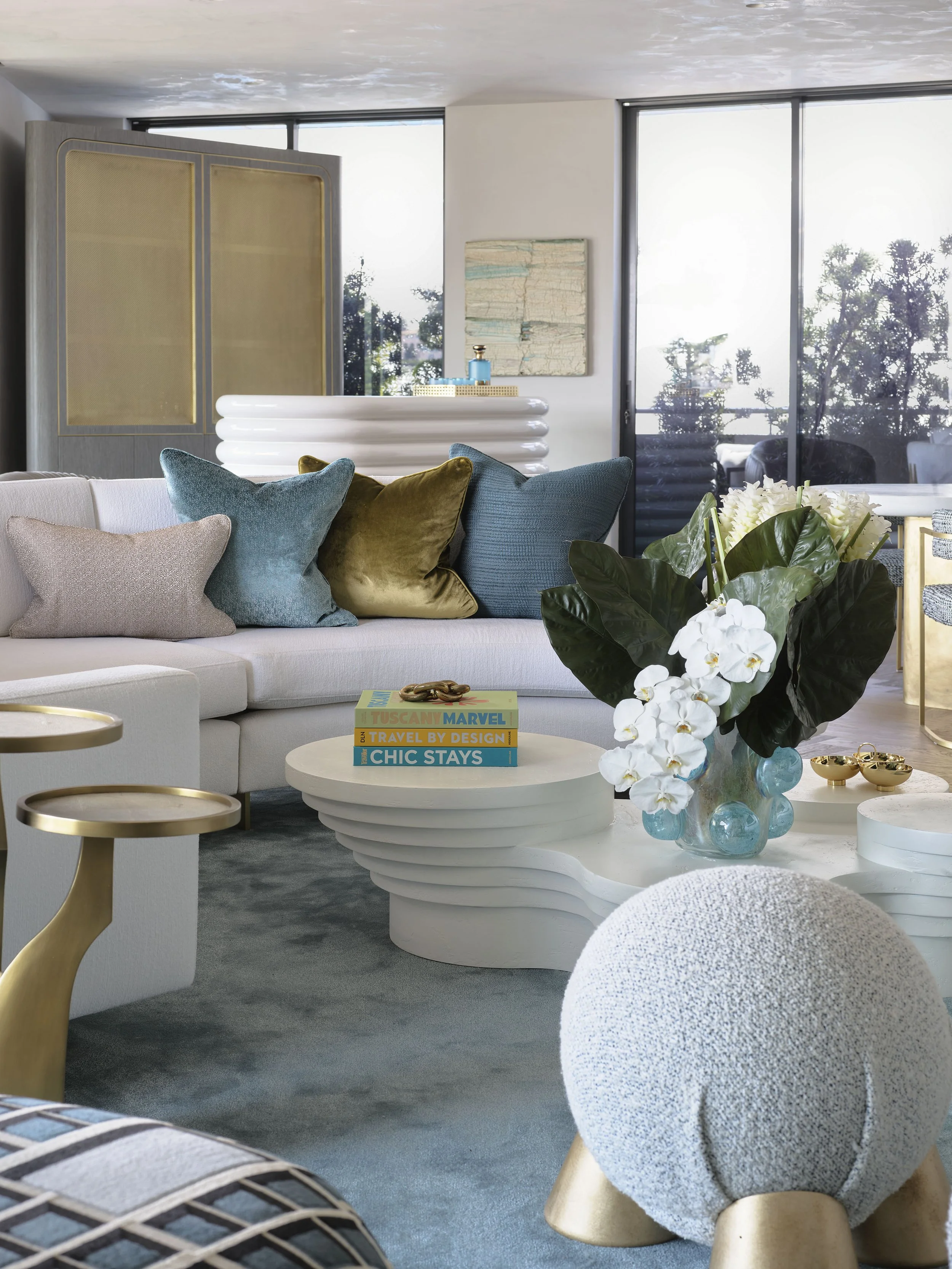 Modern living room with a white sofa, colorful velvet pillows, glass coffee table with books and flowers, and a textured ottoman.