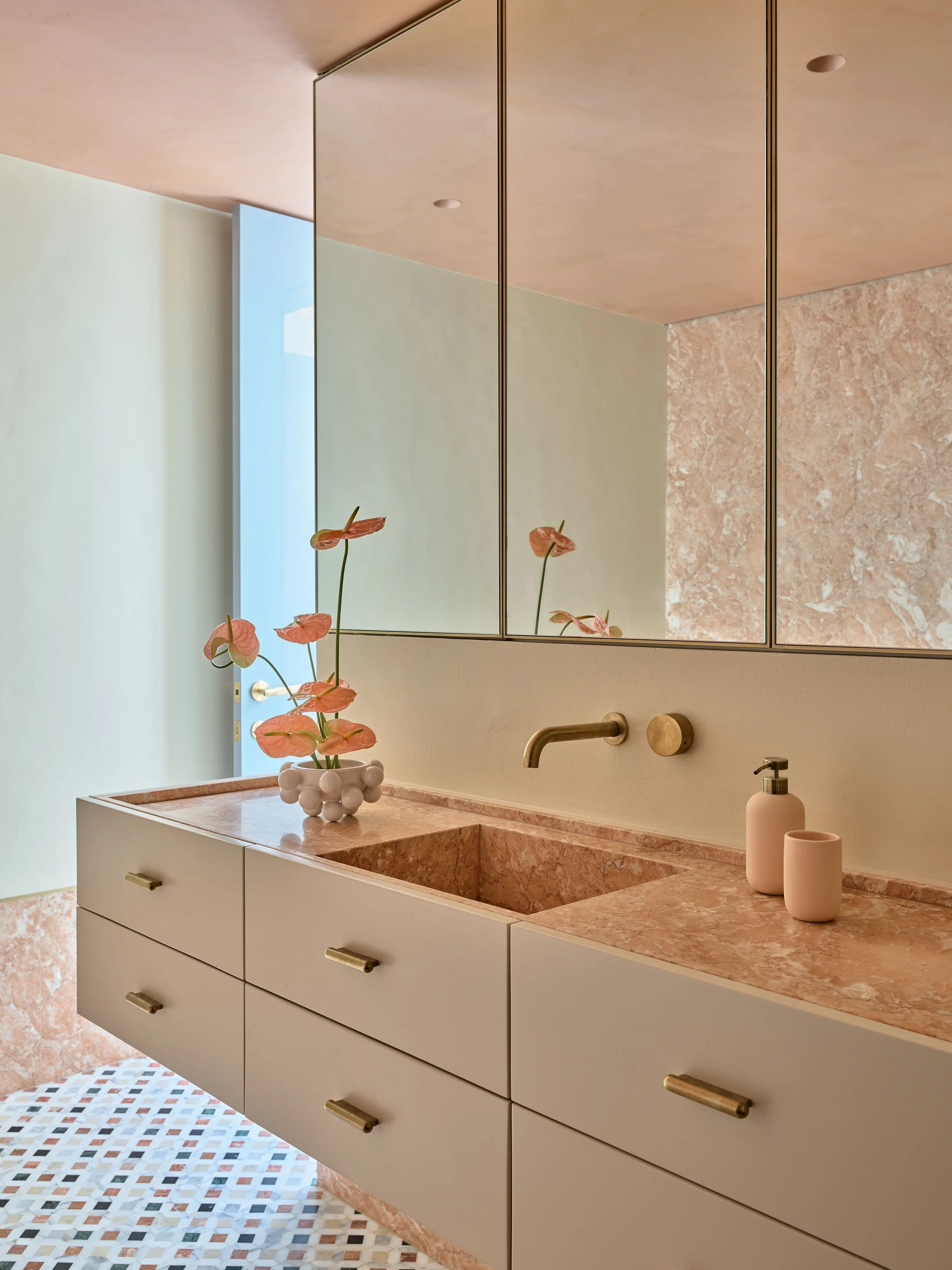 Modern bathroom with a pink stone sink and countertop, a large mirror, beige cabinets with gold handles, decorative pink vase with flowers, and a patterned tile floor.