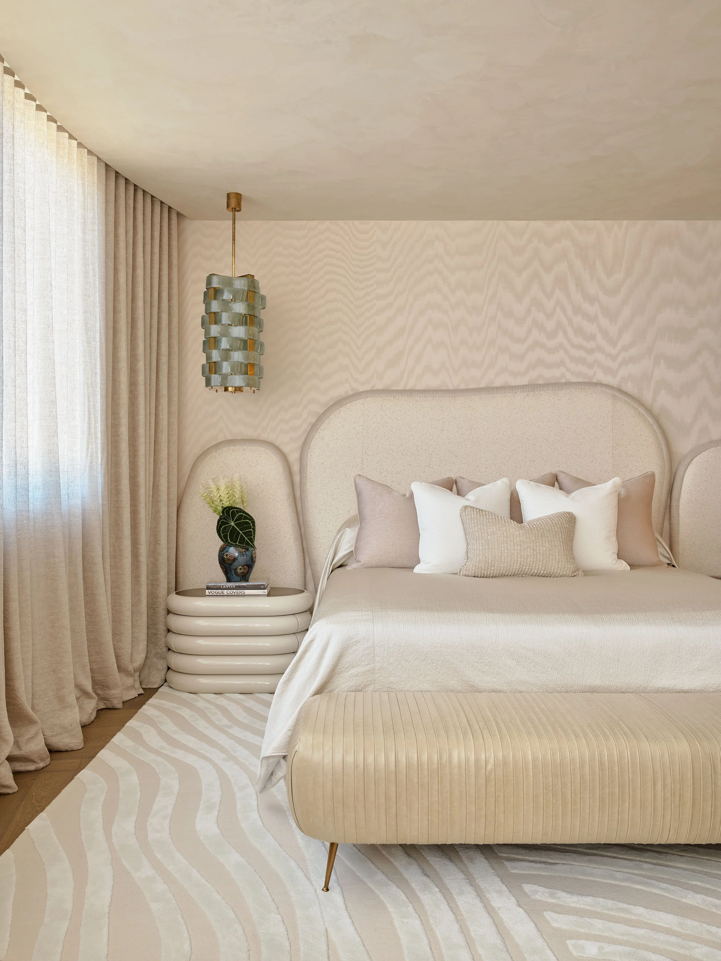 A bedroom with a beige upholstered bed, multiple pillows, a tufted bench at the foot, and a round side table with a vase and a flower, with beige curtains and a pendant light.