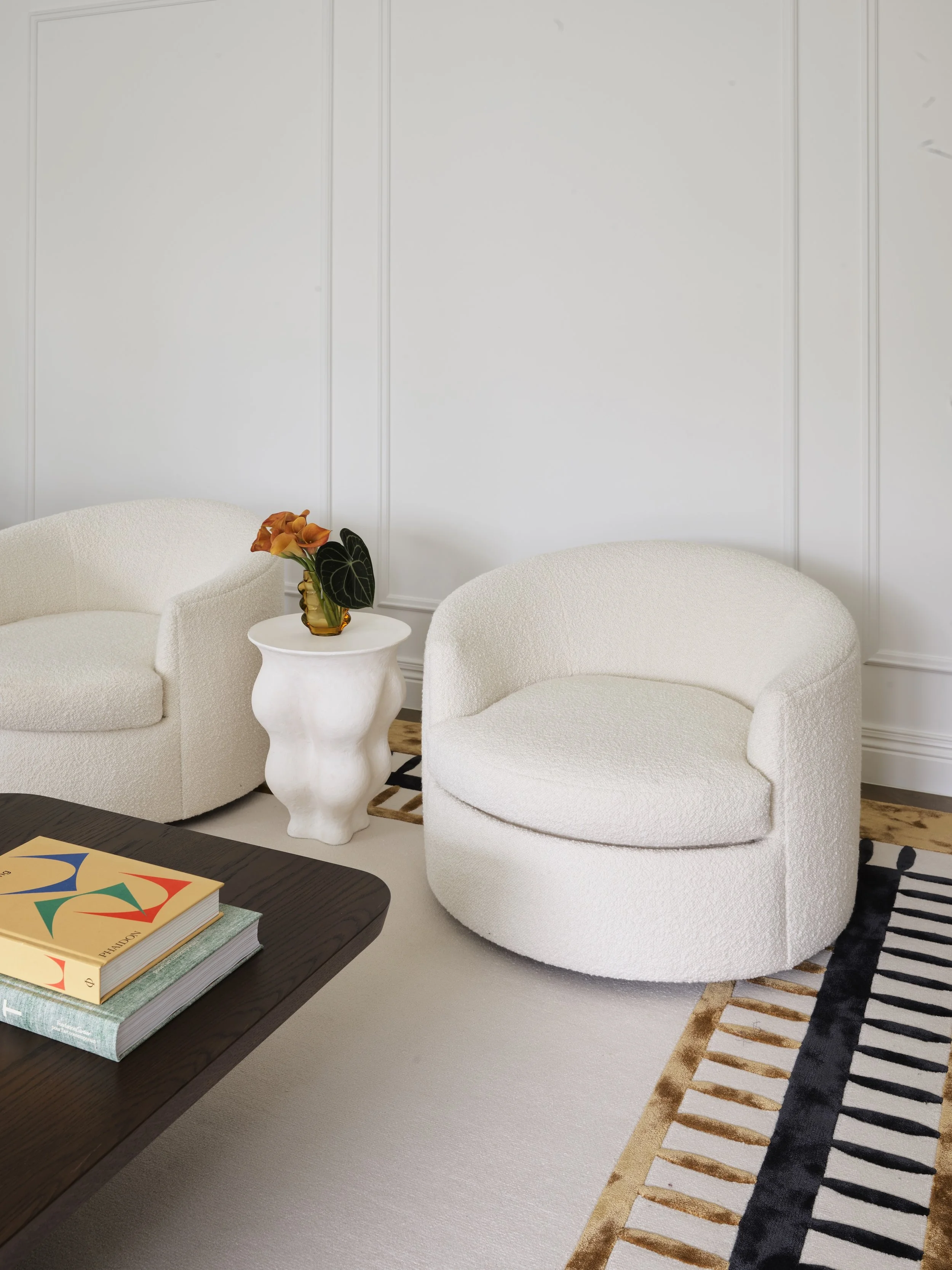Living room with white textured armchairs, a white side table with a potted plant, a dark wooden coffee table with books, and a patterned rug, against white paneled walls.