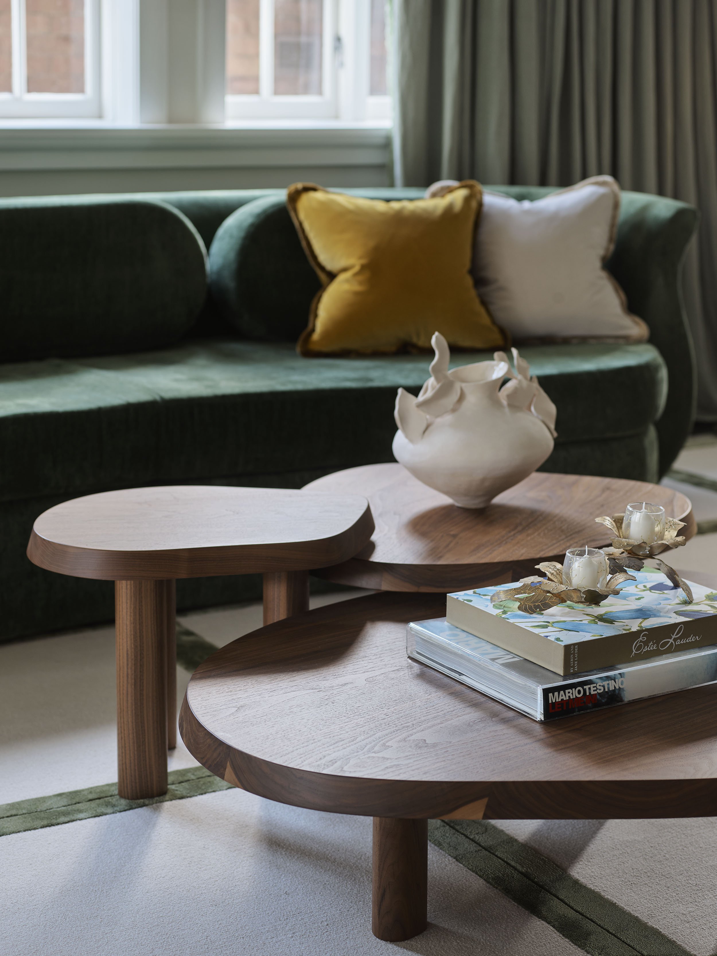 Living room with a dark green velvet sofa, yellow and white pillows, two nested wooden coffee tables, a white ceramic vase, hardcover books with decorative candle holders, and a window with curtains in the background.