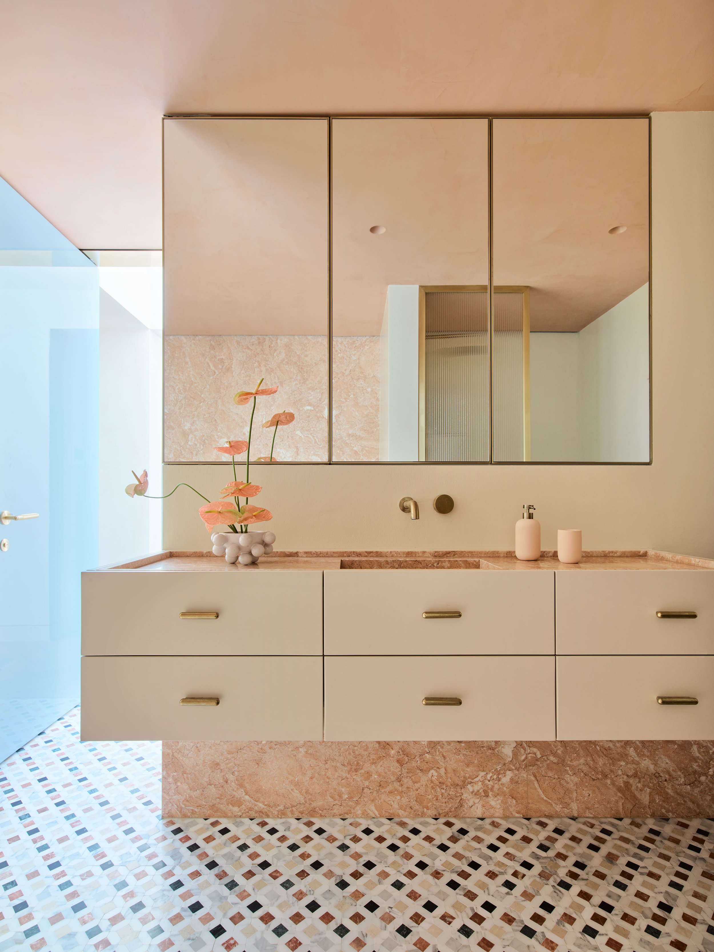 Stylish bathroom vanity with large mirror, pink marble countertop, white cabinets with gold handles, pink flowers in a white vase, and ceramic soap dispensers. Tiled floor with geometric pattern.