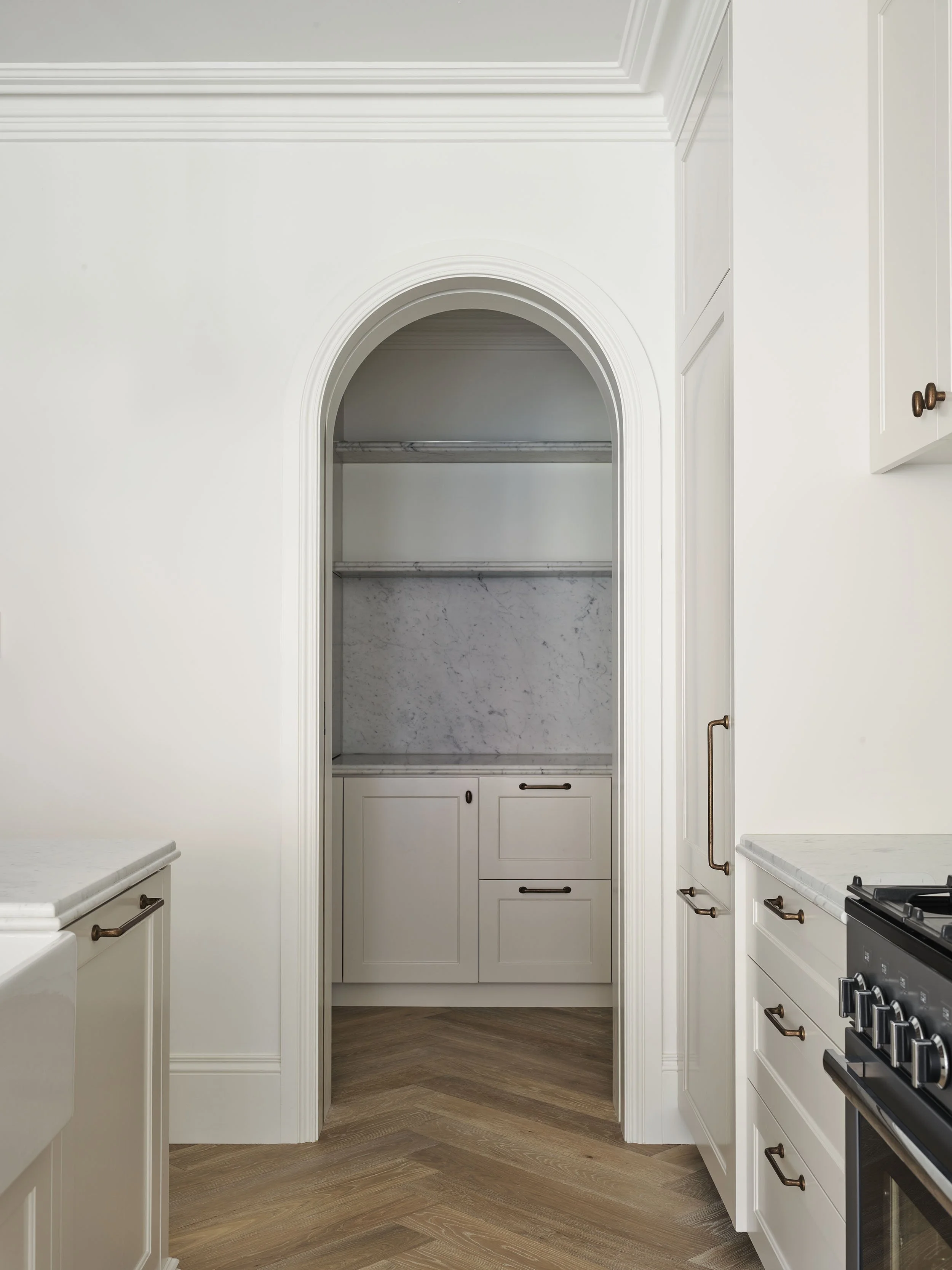 A kitchen with white cabinets and a marble countertop, a gas stove on the right, and a narrow arched entryway leading to a small nook with marble shelves and cabinets.