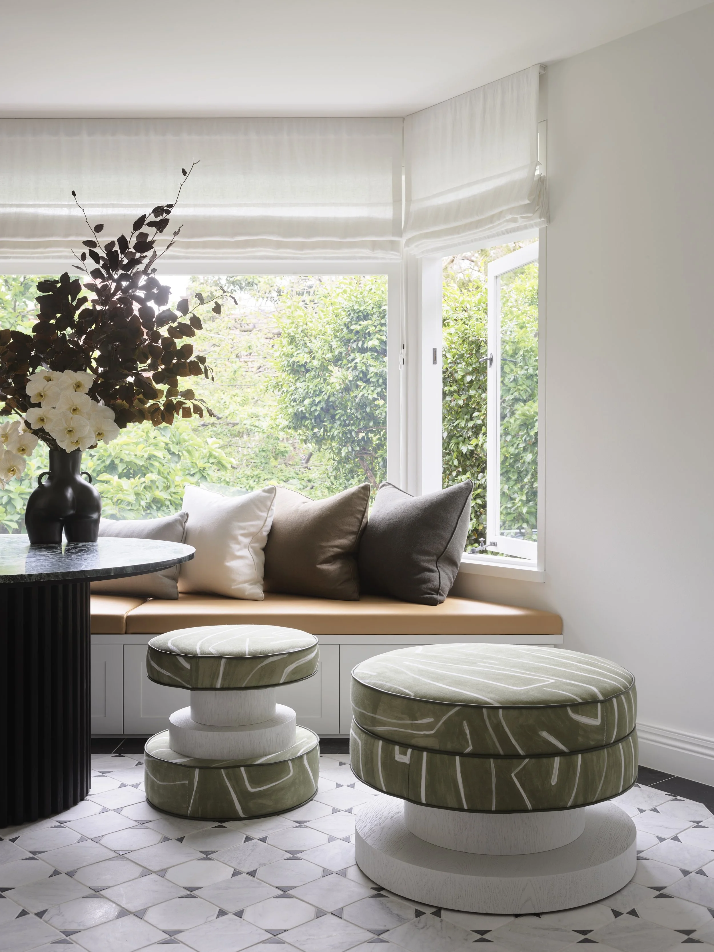 Bright sitting area with window seat, beige and brown cushions, a black vase with white flowers, and patterned ottomans on a white and black tiled floor.