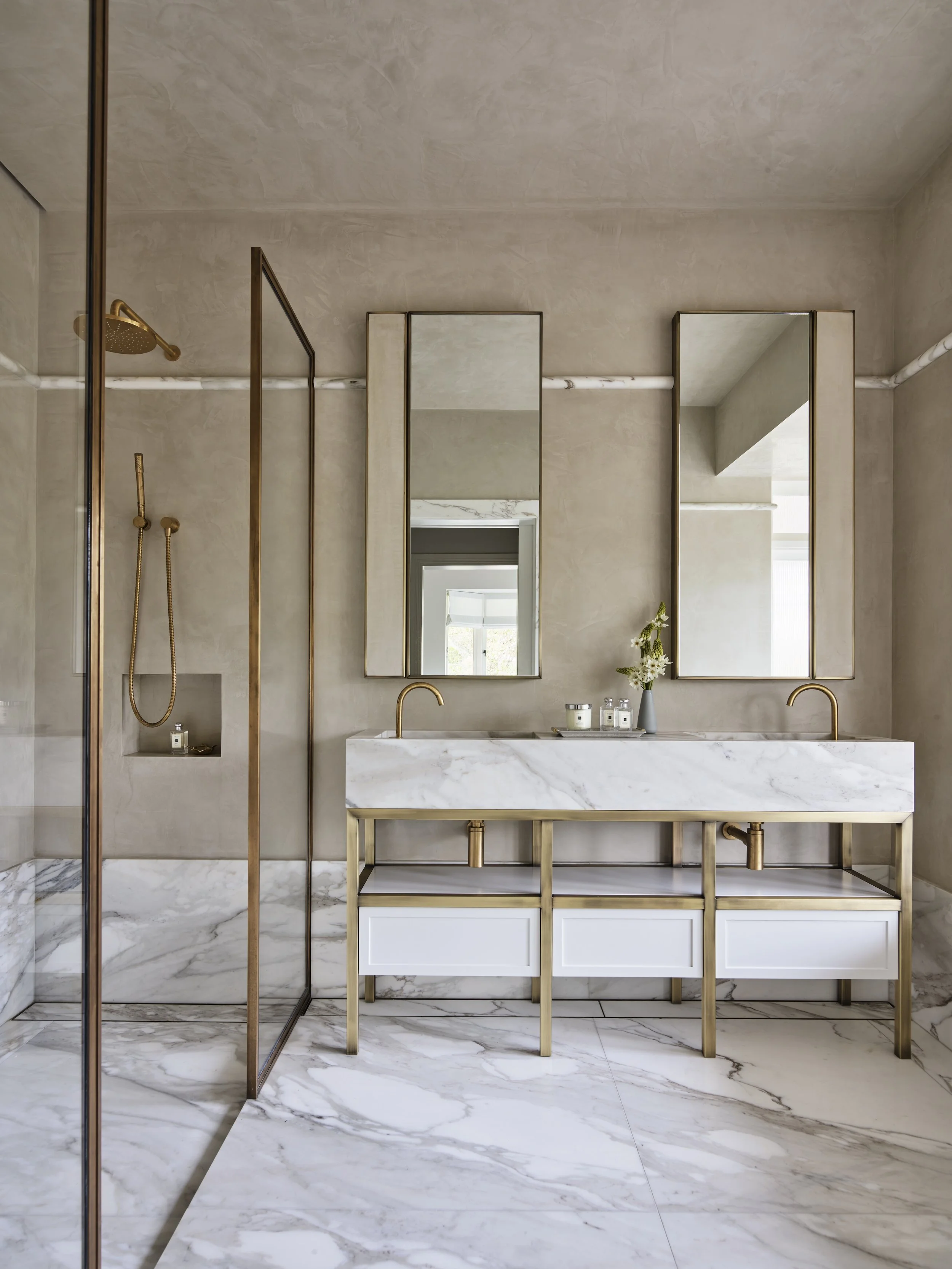 Modern bathroom with dual marble sinks, gold fixtures, and large mirrors, featuring a walk-in shower with a gold showerhead and handheld sprayer.