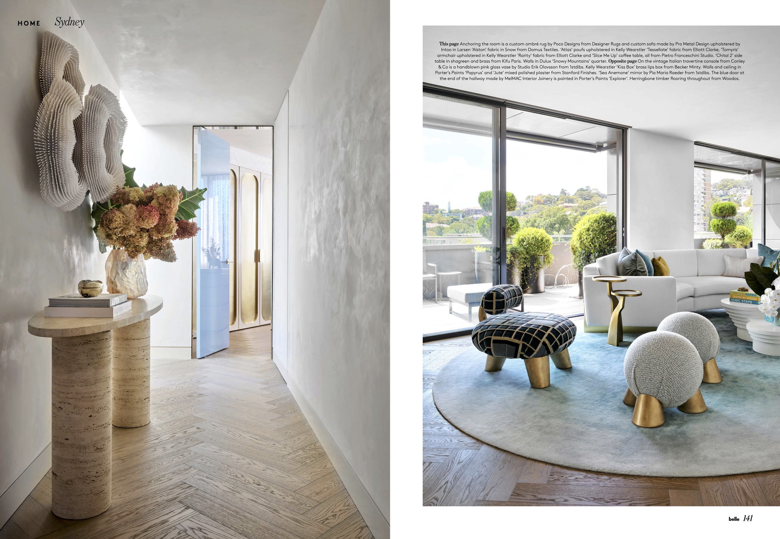 Interior of a modern living space with a hallway featuring a floral arrangement on a curved stone table, textured white walls, and light wooden flooring. In the adjacent room, there is a spacious lounge with large windows, green plants, a white sectional sofa, patterned stools, and a blue rug.