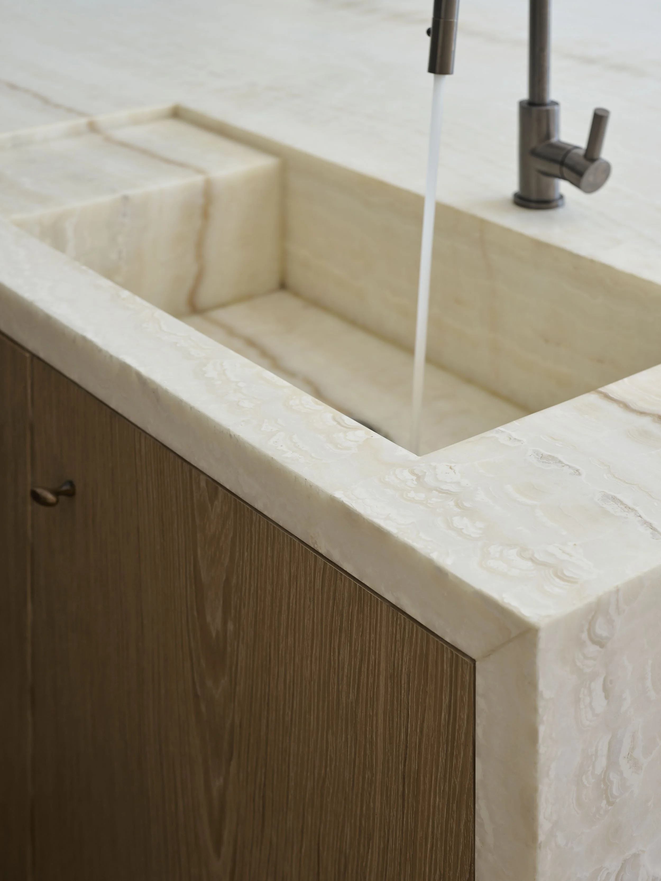 Close-up of a beige marble kitchen sink with a modern metallic faucet and running water, installed on a wooden cabinet.