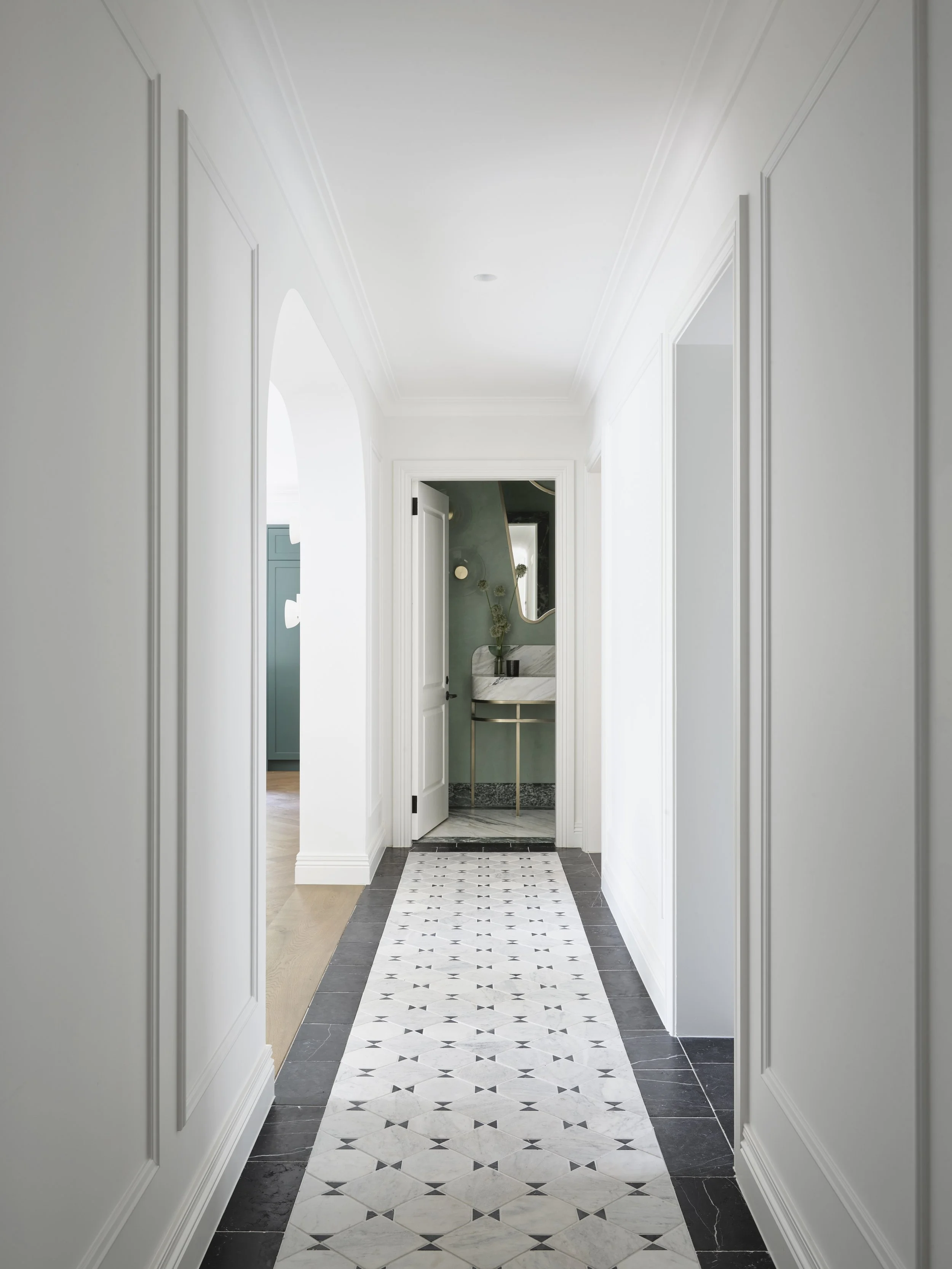 A bright, modern hallway with white walls and intricate crown molding, leading to a bathroom with a marble sink, gold accents, and a mirror. The floor features a geometric black and white tile pattern.