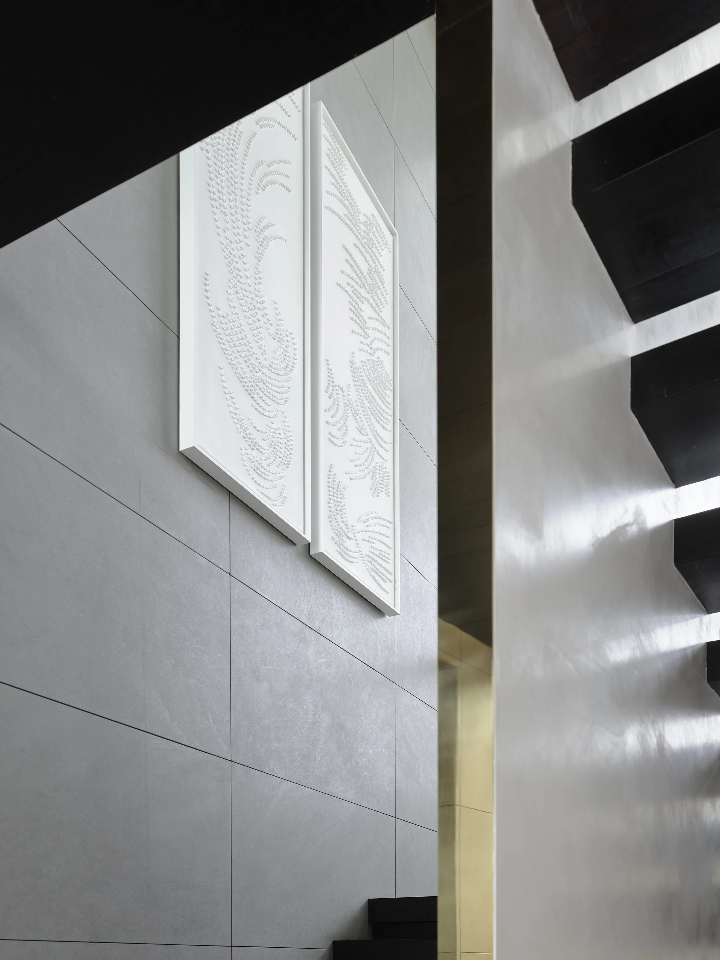 Interior view of a modern building with gray tiled walls, black staircase, a gold column, and a white textured art piece on the wall.