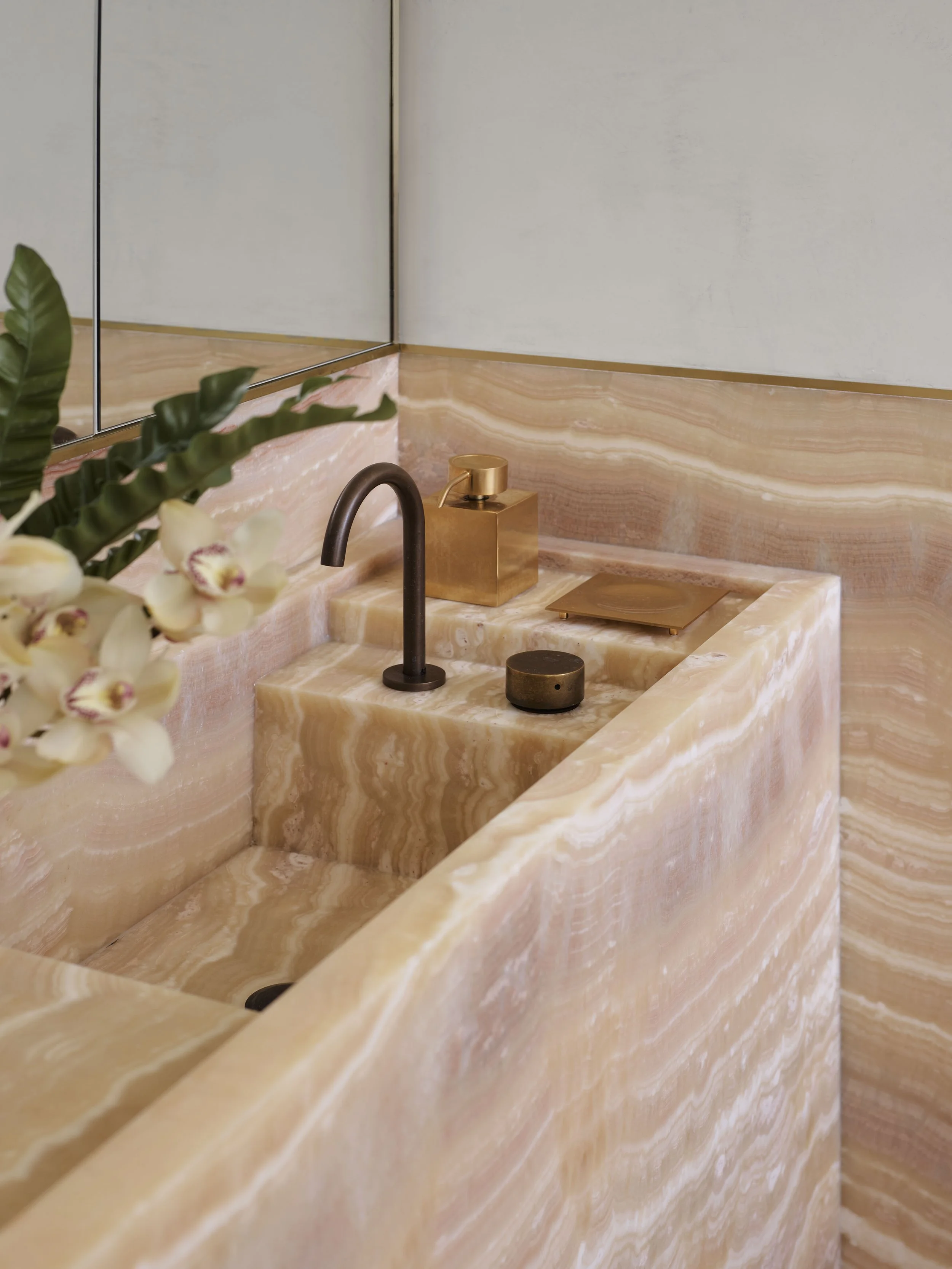 Close-up of a modern bathroom sink with pink and white marble finish, gold soap dispenser, black faucet, decorative black container, and gold tray, with a mirror and green plant with white orchids in the background.
