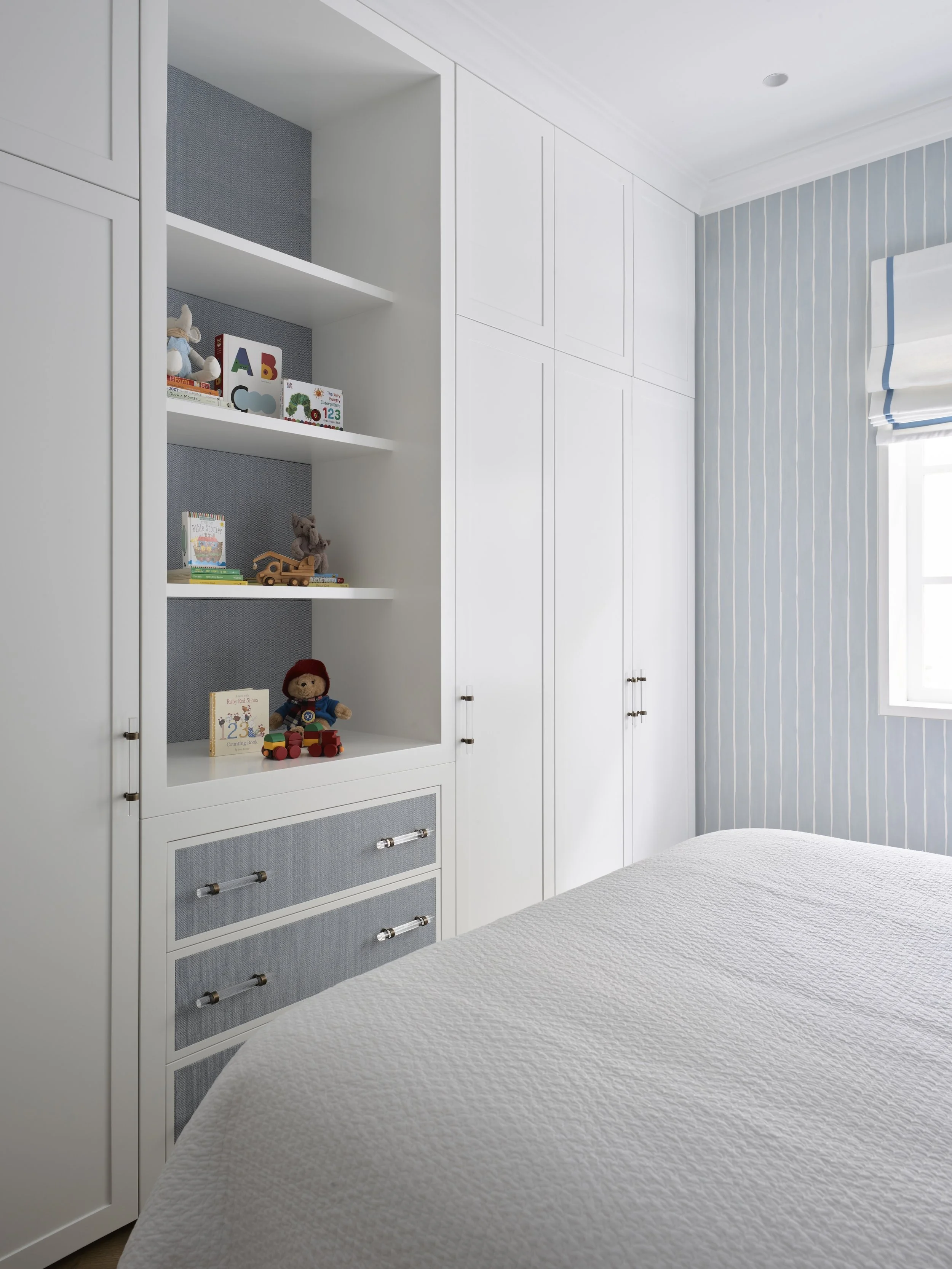 A white bedroom with built-in cabinets and a window with blue and white striped curtains. A bed with a white textured cover is in the foreground, and shelves with children's toys and books are visible on the left.