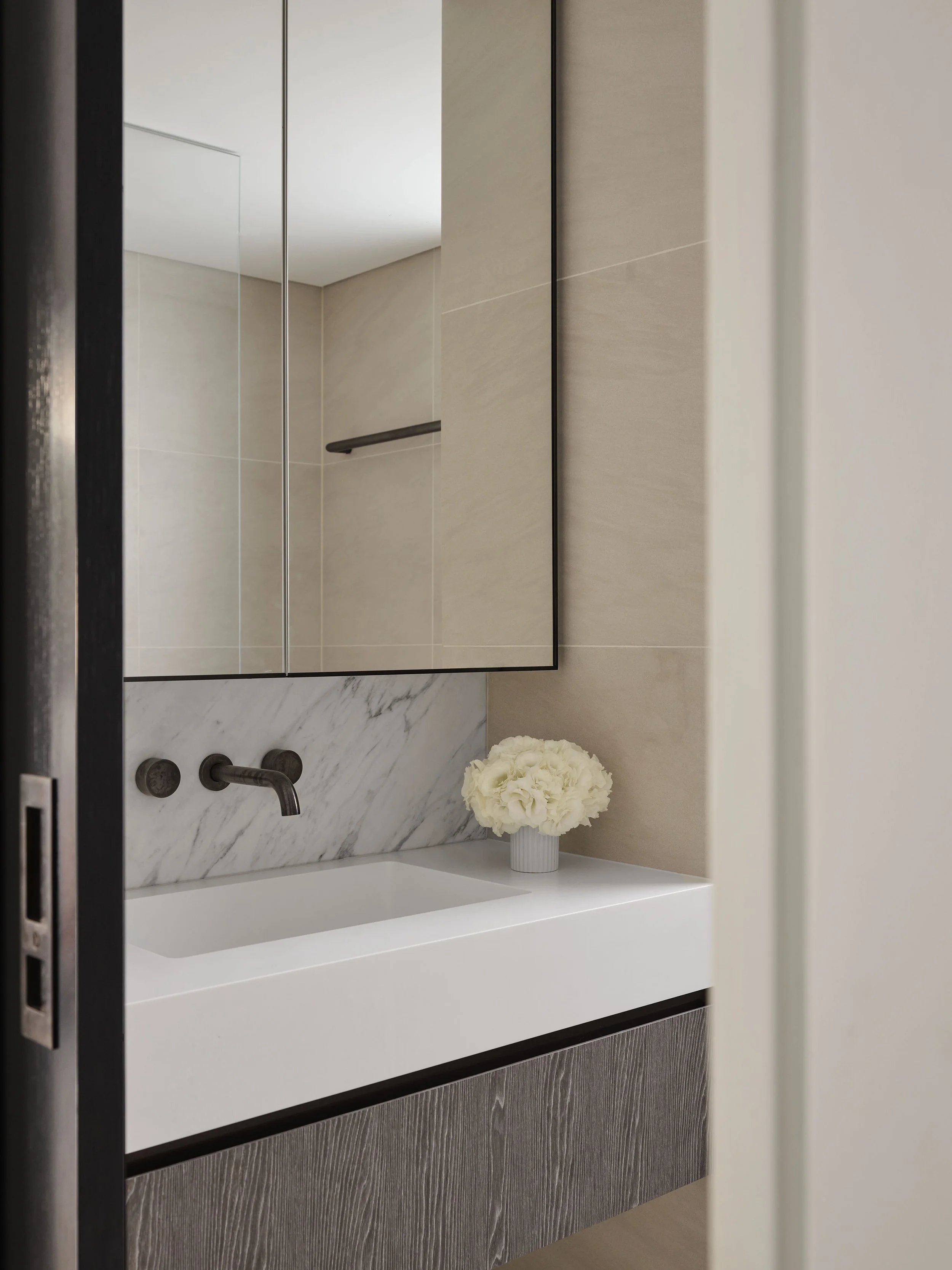Modern bathroom with a white rectangular sink, mounted on a wood-textured vanity, a marble backsplash, and a large mirror. White flowers in a gray vase sit on the countertop.