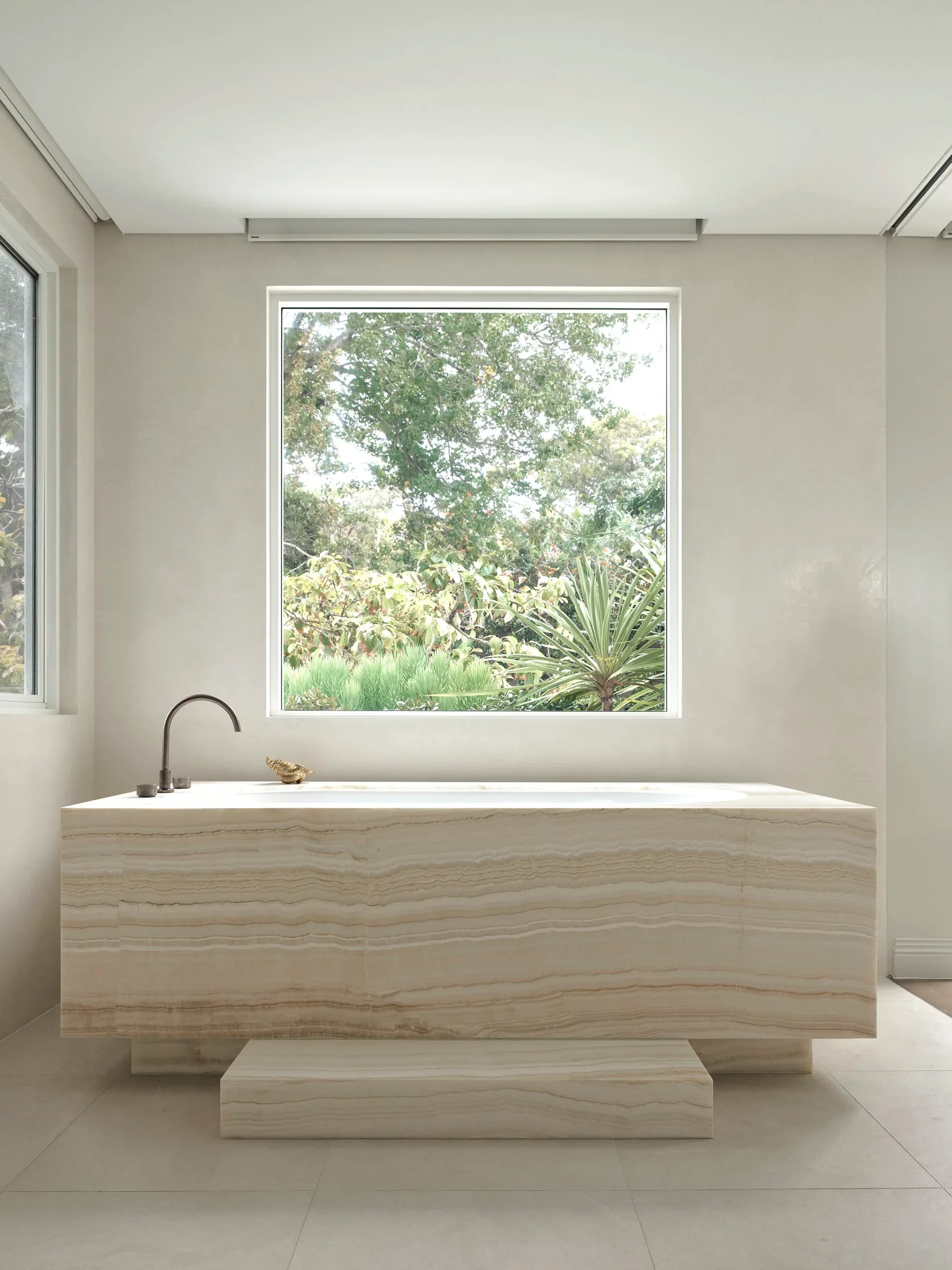 Modern bathroom with a freestanding beige marble bathtub, a silver faucet, and a large window overlooking greenery.