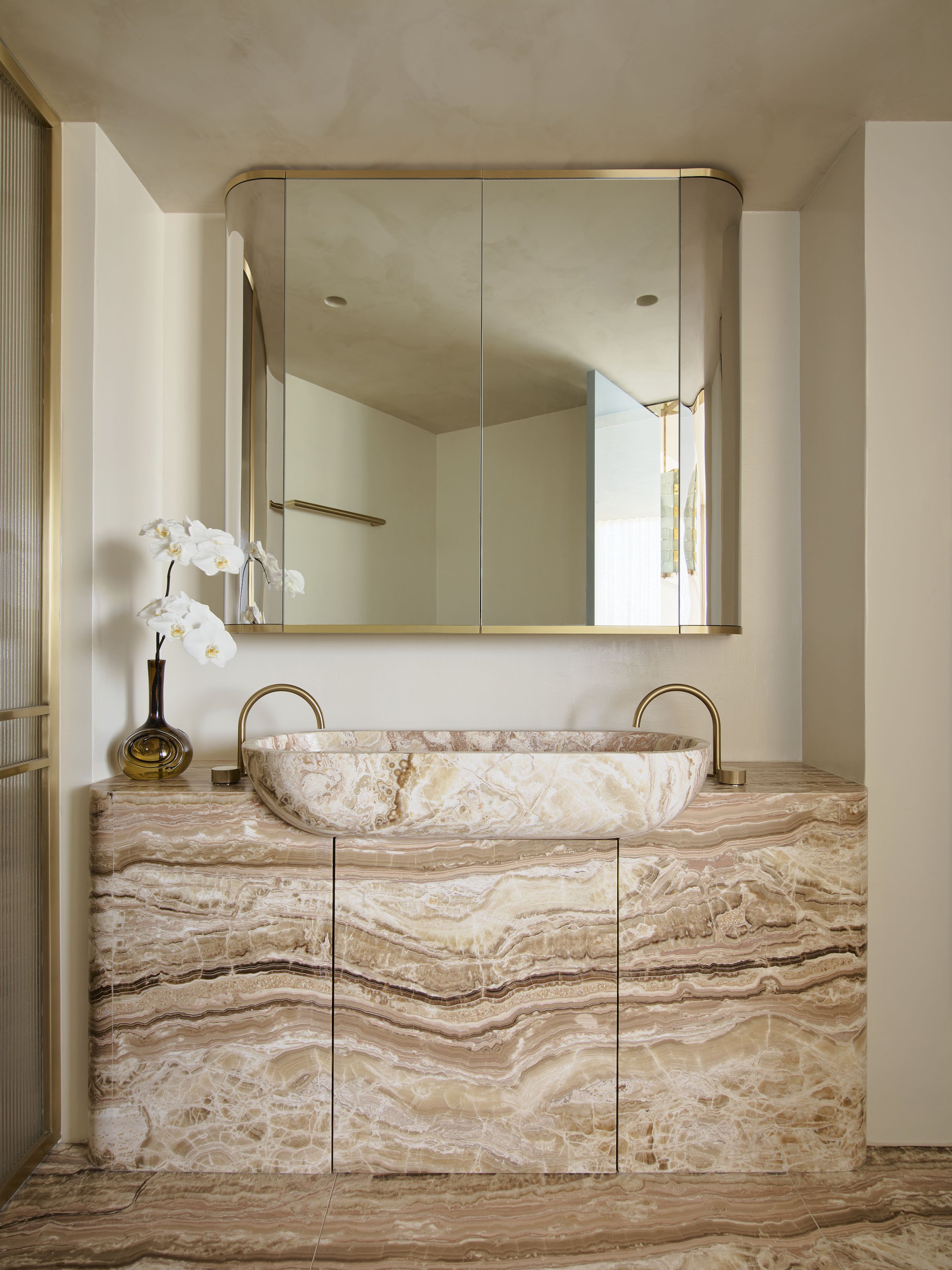 A bathroom vanity with a marble countertop, a rounded marble vessel sink, a large mirrored cabinet, a small decorative vase with white orchids, and gold fixtures.