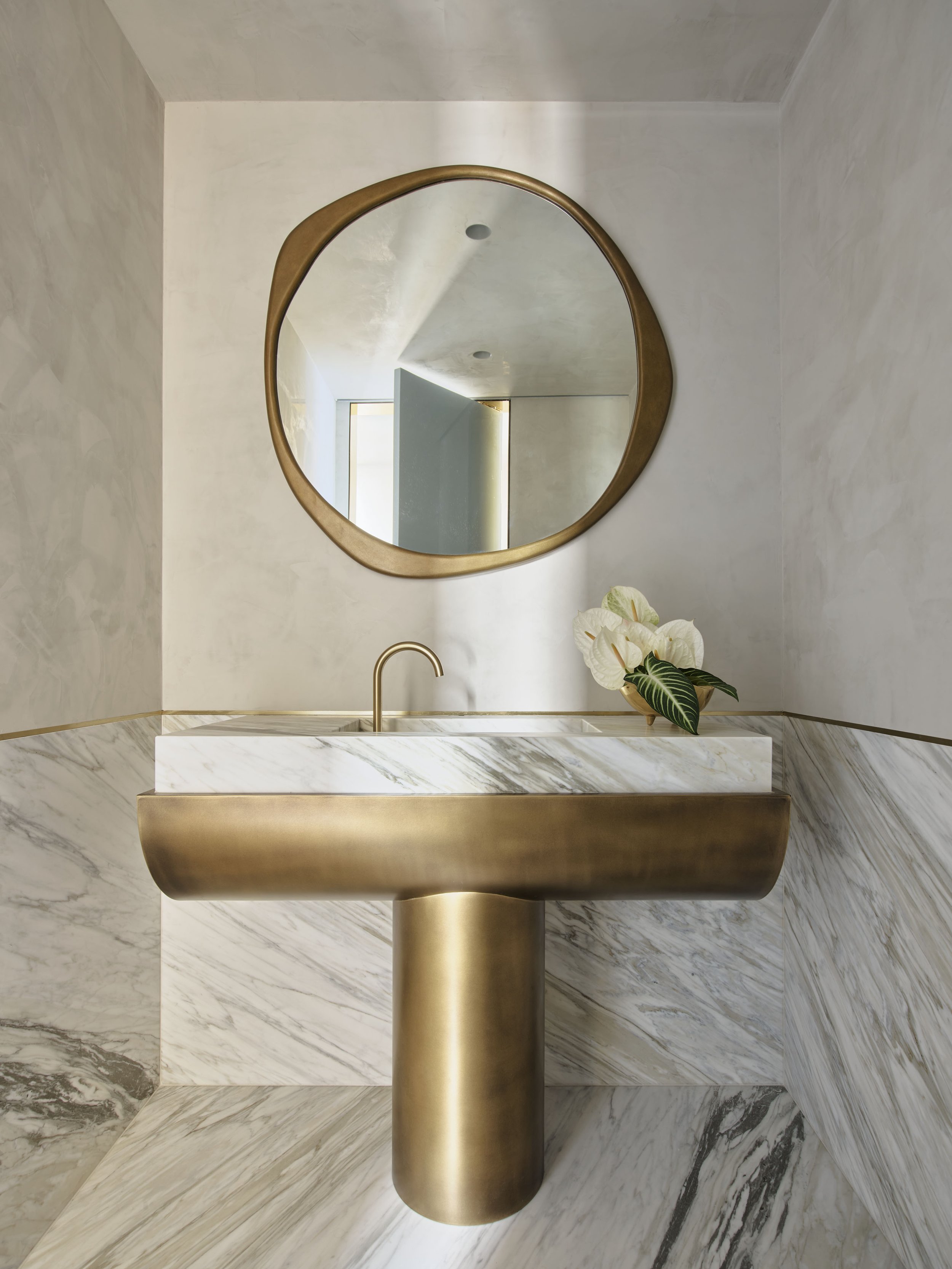 A modern bathroom vanity with a white marble sink, a round mirror with a gold frame, and a small plant with white flowers and green leaves.