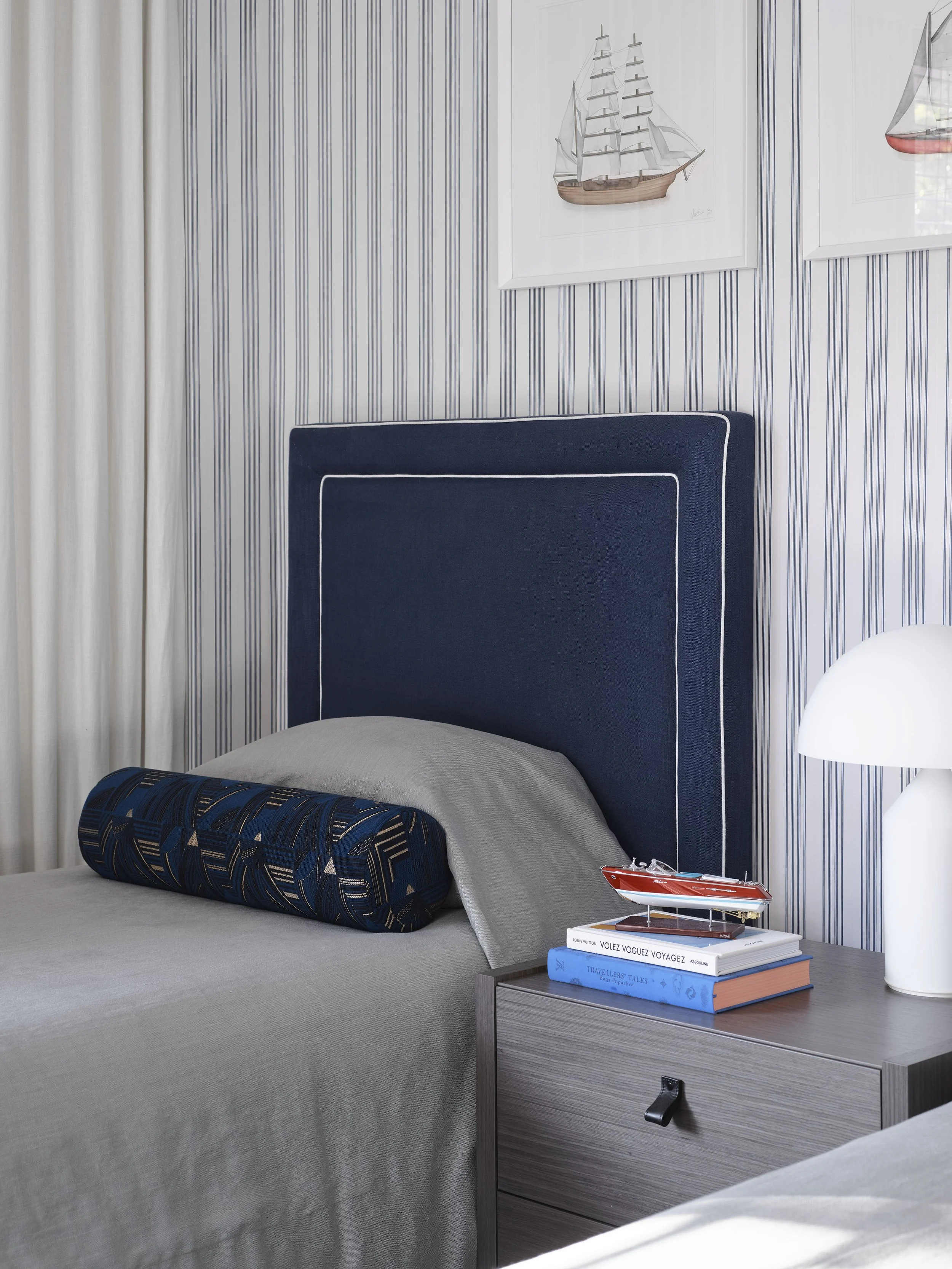 A bedroom with a navy blue upholstered headboard against striped wallpaper, a small bedside table with a model ship, books, and a lamp.