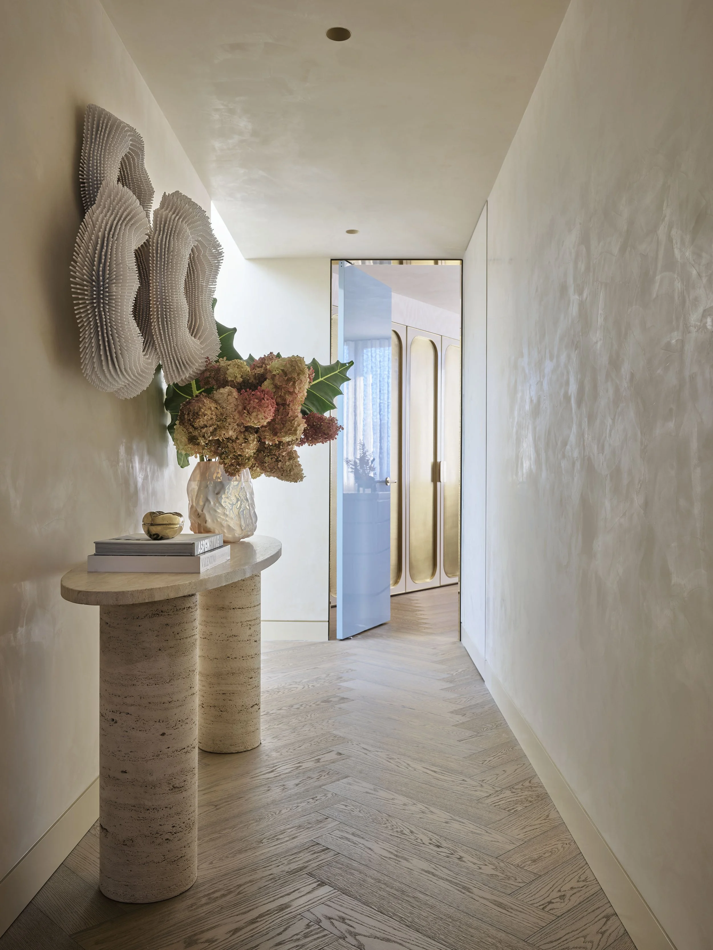 Interior hallway with beige walls, light wood flooring, a marble console table with a vase of pink flowers and books, decorative wall art, and a partially open door leading to a room with gold accents.