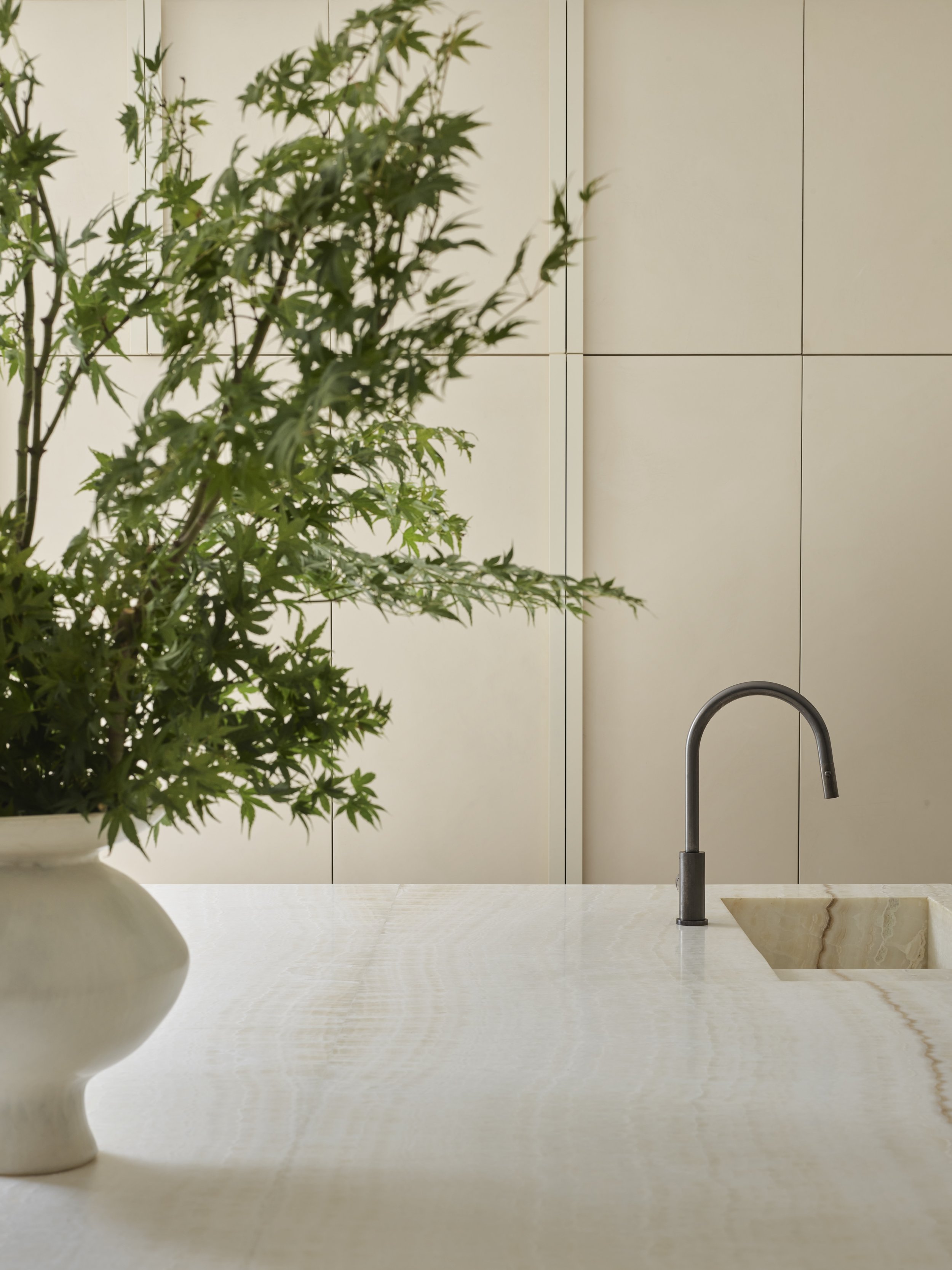 A kitchen countertop with a potted plant and a modern sink with a curved faucet, against a minimal beige tiled wall.