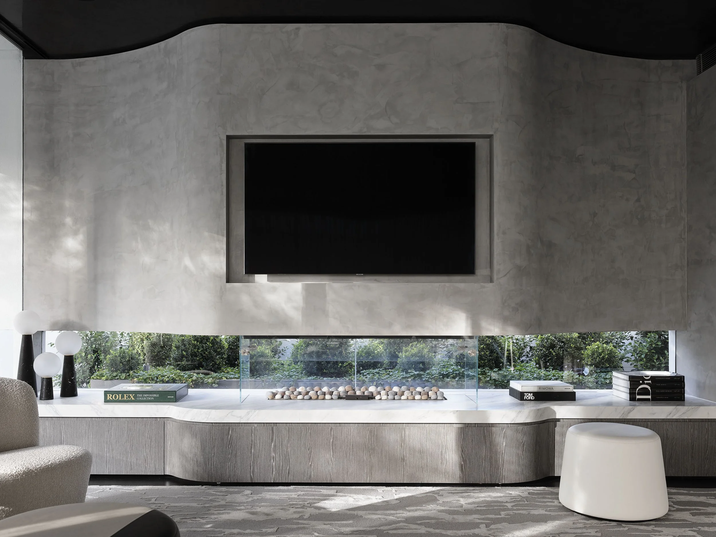 Modern living room with a large wall-mounted flat-screen TV, decorative objects including black vases with white globes, a book titled 'ROLEX,' stacks of books, and a glass-enclosed arrangement of seashells on a sleek marble entertainment center, wit