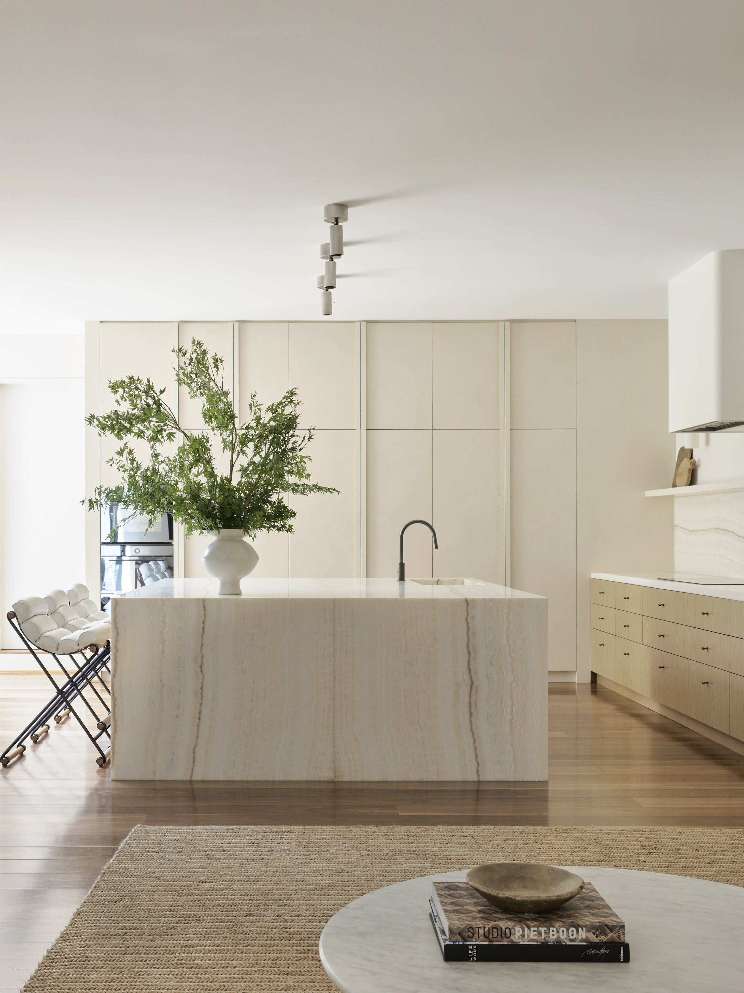 Minimalist kitchen with a large white marble island, a white vase with green foliage, and light wood cabinetry, with a beige rug and round coffee table in the foreground.