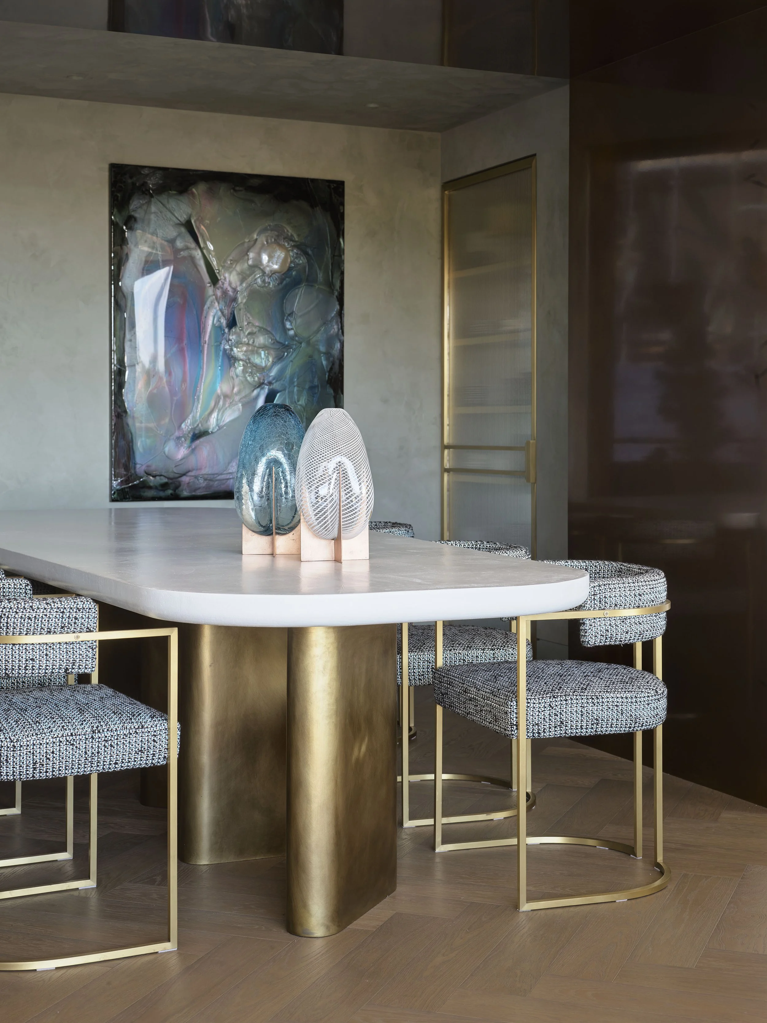 A modern dining room with a white oval table, four chairs with black and white checked upholstery and gold metal frames, two decorative glass sculptures on the table, a large abstract painting on the wall, and a gold-framed glass door.