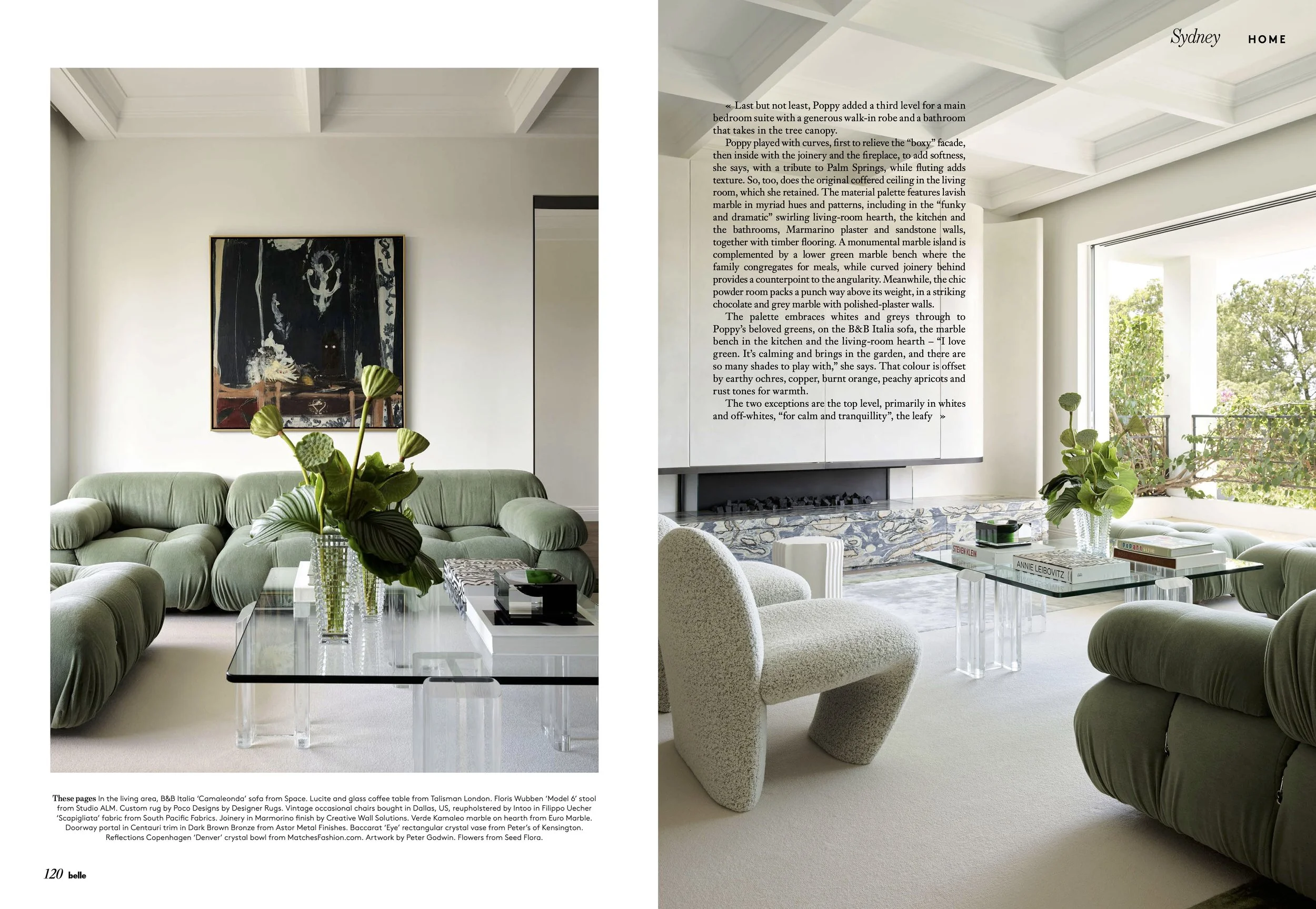 A living room with green tufted sofas, a glass coffee table with vases and books, beige textured chair, decorative plants, white walls, a large abstract painting, and a fireplace with a marble hearth and a large window with greenery outside.