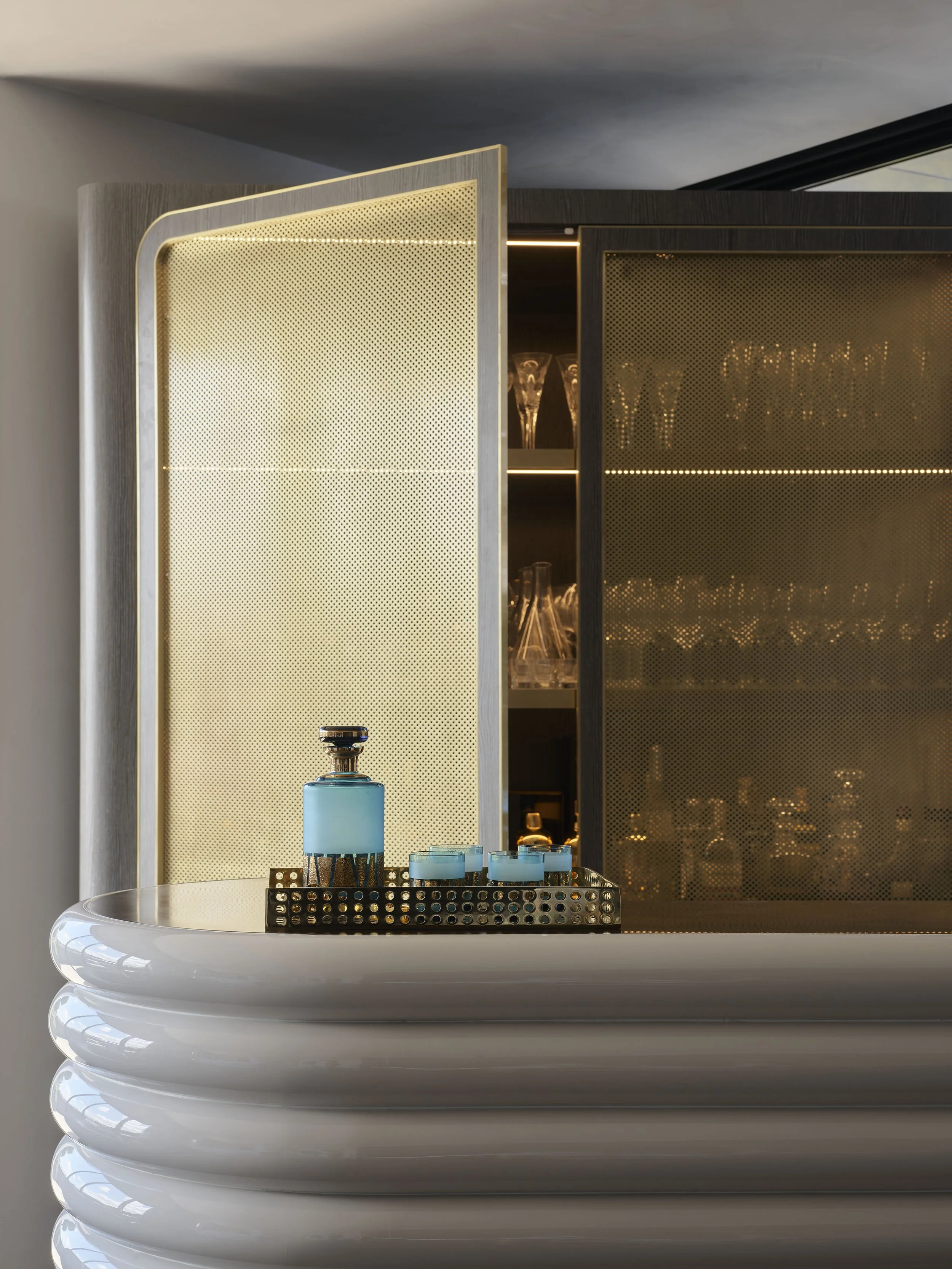 Close-up of a modern bar with a gold-toned, perforated partition, glassware inside, and a tray with blue candles and a blue decanter on a glossy, white, ribbed countertop.