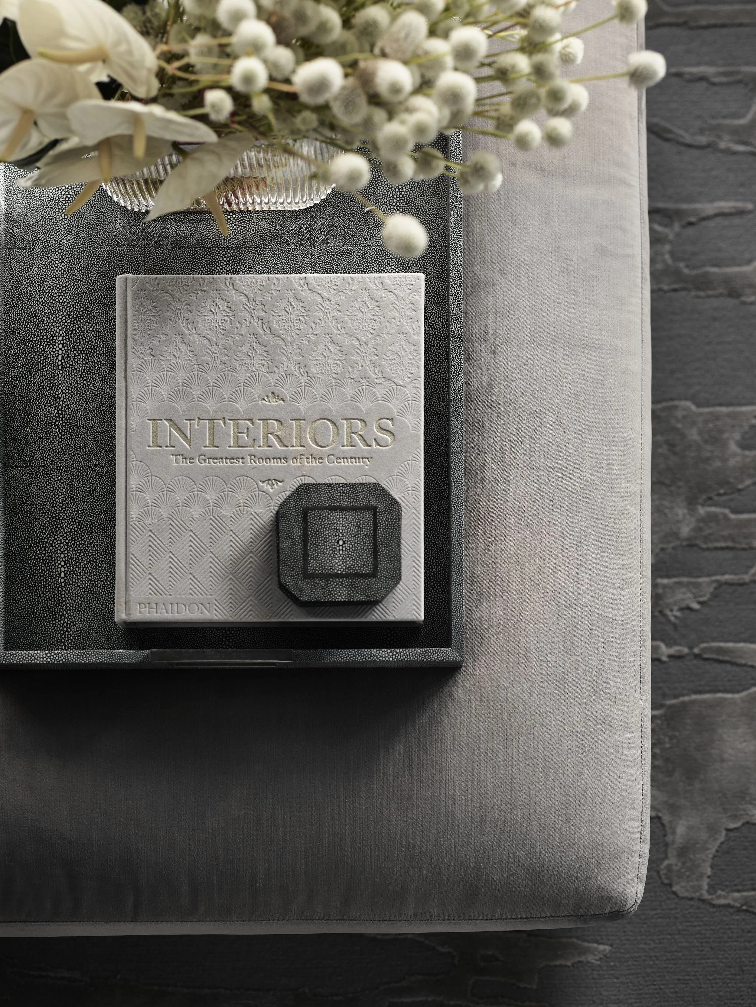 A decorative tray with a textured book titled 'INTERIORS: The Greatest Rooms of the Century' and a small black box, alongside a floral arrangement of white flowers and green leaves, set on a gray fabric-covered table against a stone wall.