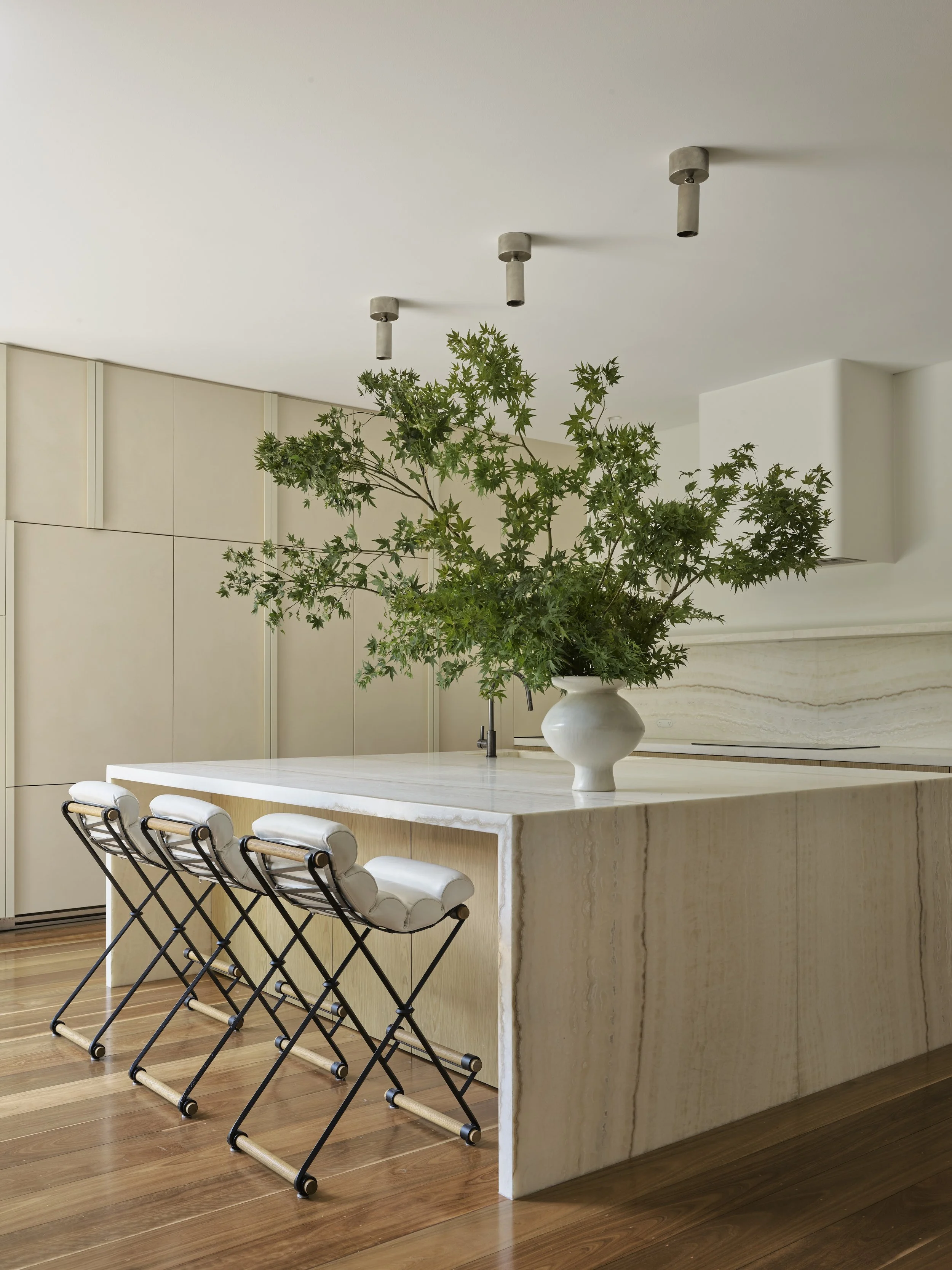 Modern kitchen with a large marble island, white bar stools, and a large vase with green foliage as a centerpiece.
