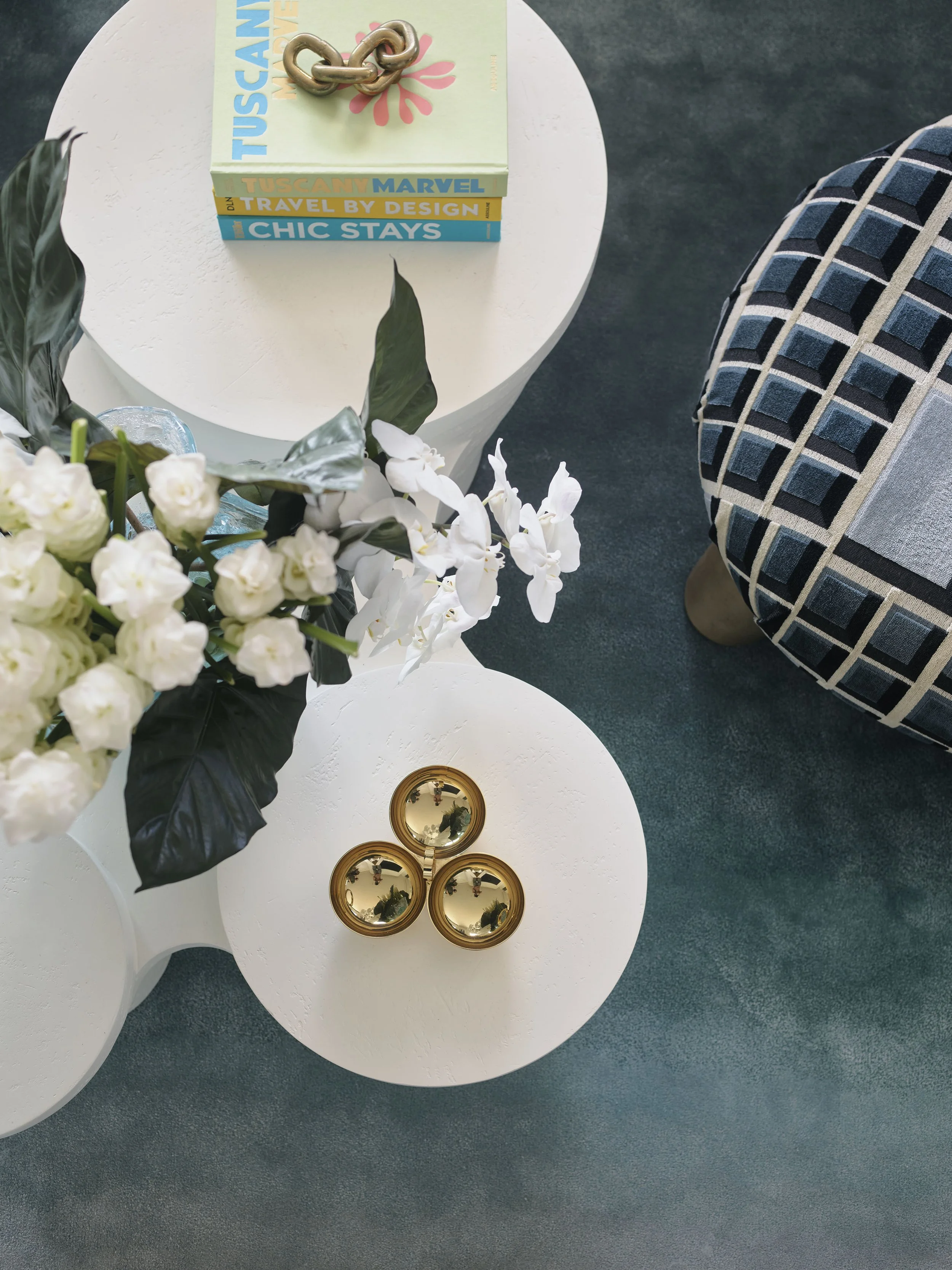 A white side table with a stack of colorful books, a decorative chain, and three small golden bowls. A vase with white flowers and green leaves is partially visible. A patterned blue and gray cushion is on the right, and a dark carpeted floor is in t