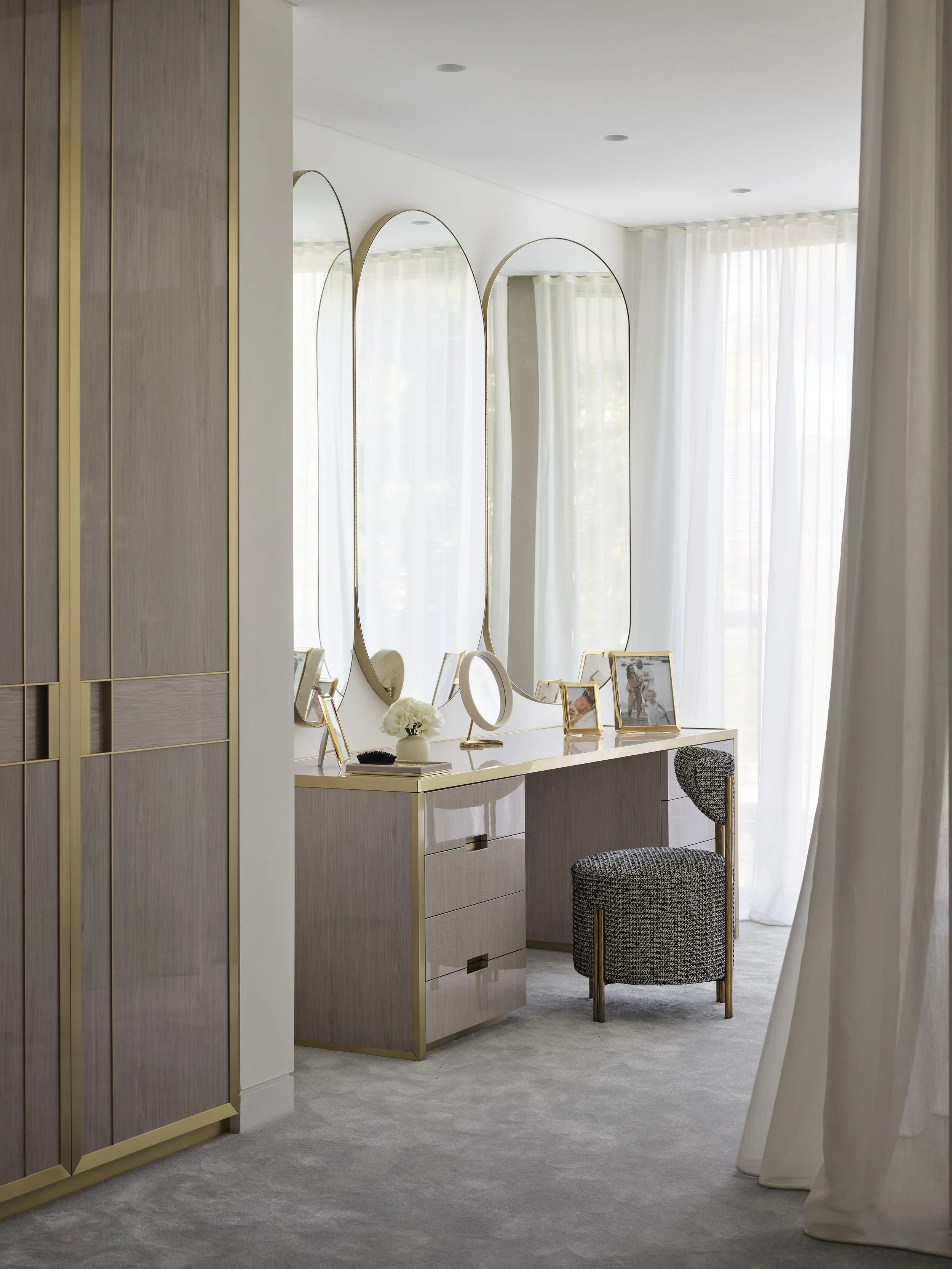 A bedroom vanity area with a light-colored wooden desk, three oval mirrors with gold frames, framed photos, a small mirror, and a flower arrangement. There's a gray upholstered stool and sheer white curtains allowing natural light to fill the room.