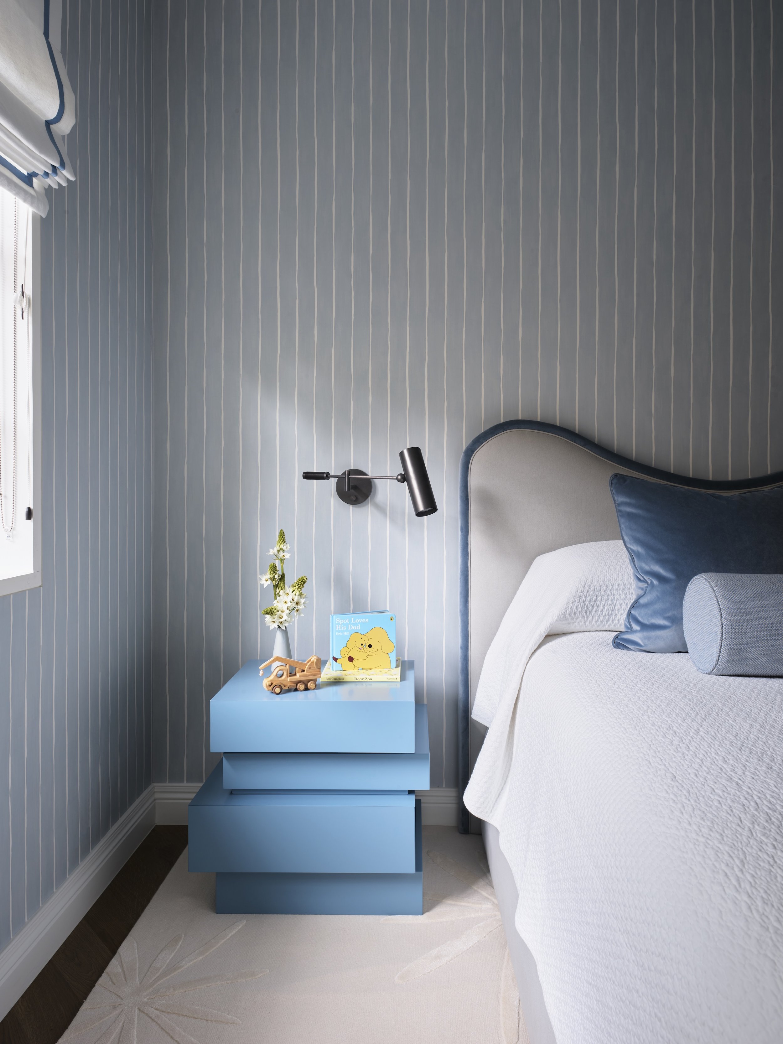 Bedroom with blue striped wallpaper, bedside table, white bed with blue pillows, black wall-mounted reading lamp, and a window with white blinds.