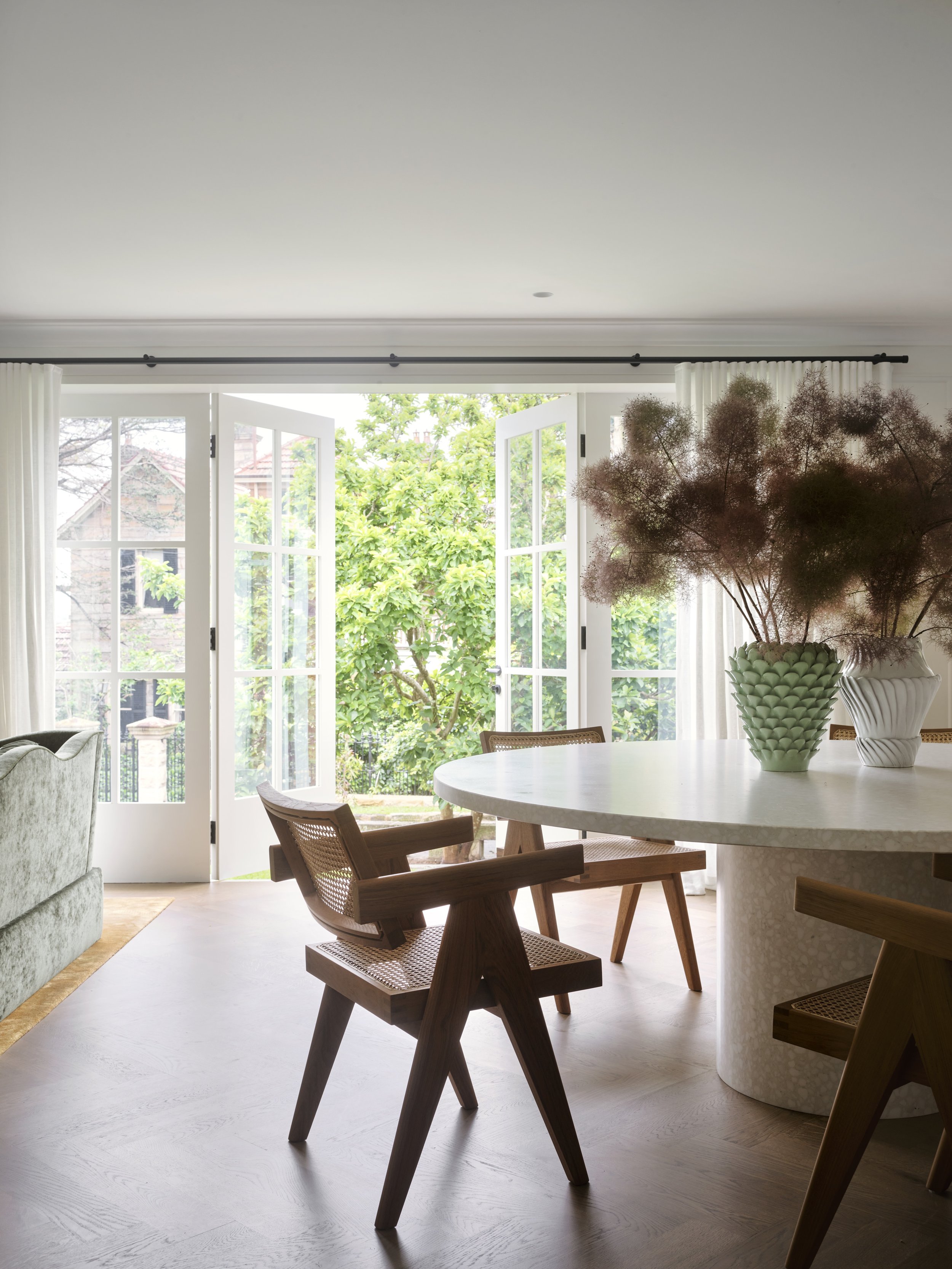 Bright living room with large open glass doors leading outside, wooden dining table with chairs, and vases with dried flowers on the table.