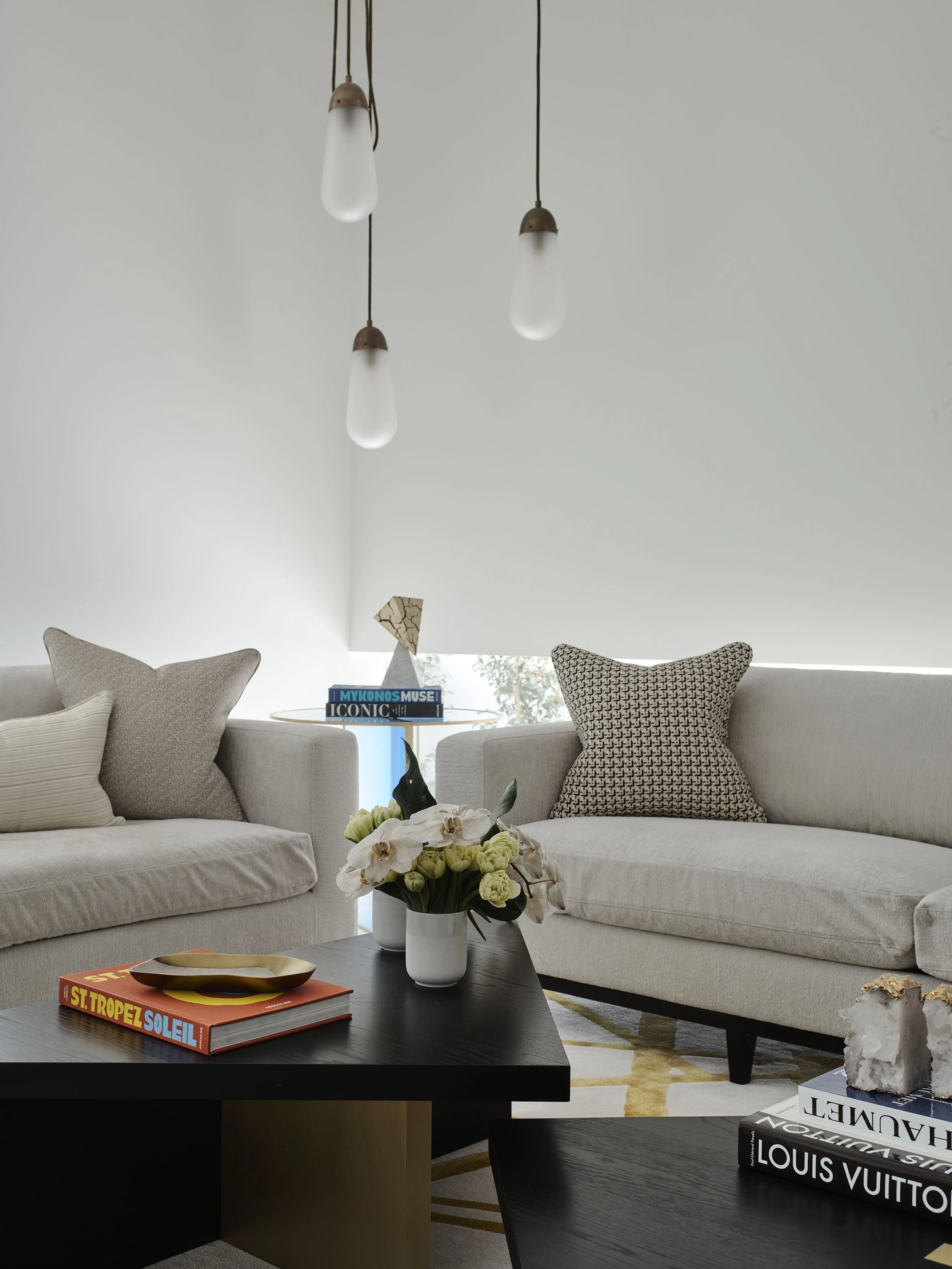 Modern living room with two beige sofas, decorative pillows, a black coffee table with books and a vase of flowers, and three hanging pendant lights.