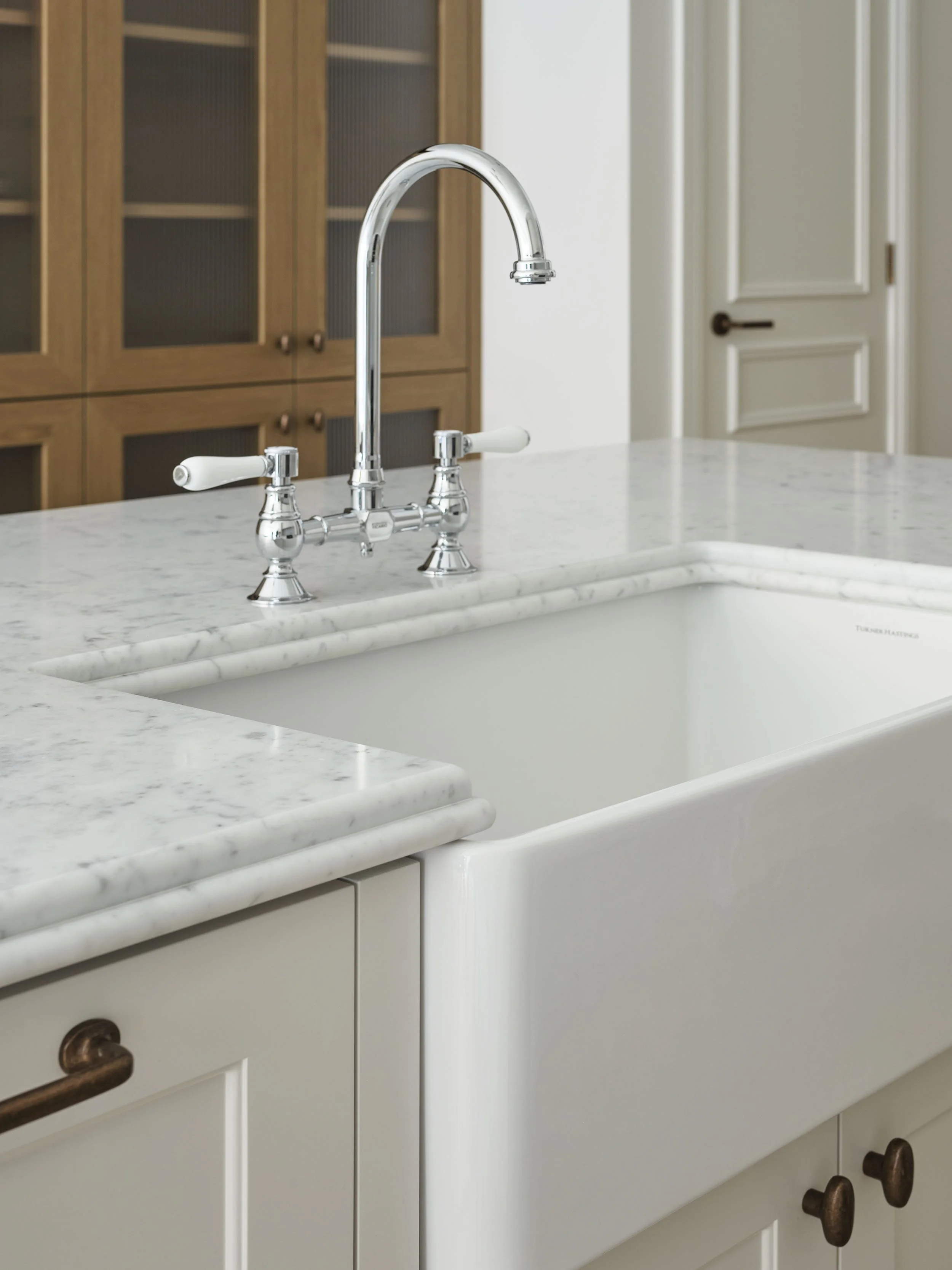 White farmhouse kitchen sink with a marble countertop and a chrome faucet, with wooden cabinets and a white door in the background.