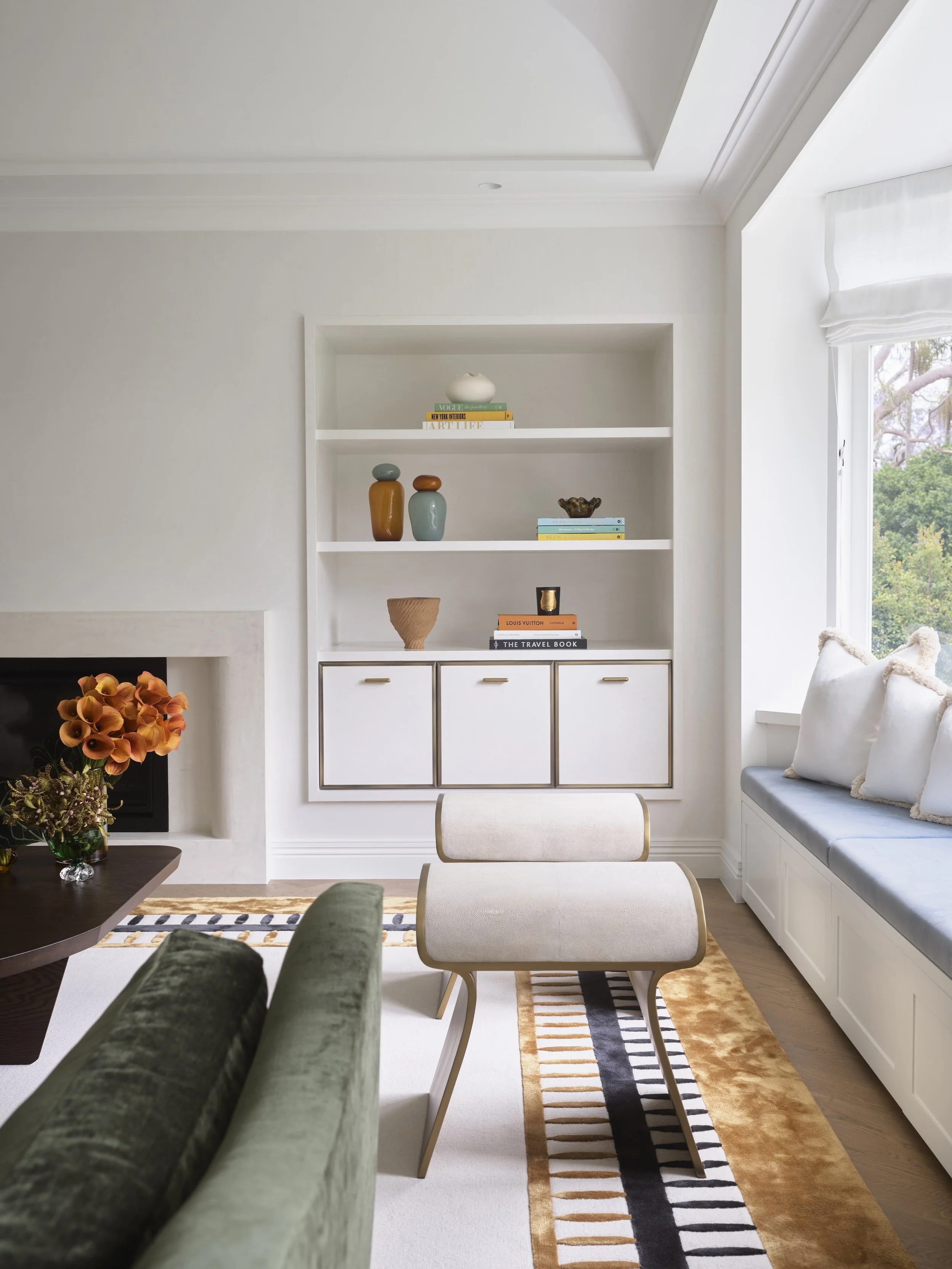 A modern living room with a built-in white bookshelf, colorful decorative vases, books, a bench by the window, a sofa, a carpet, and a coffee table with a bouquet of orange flowers.