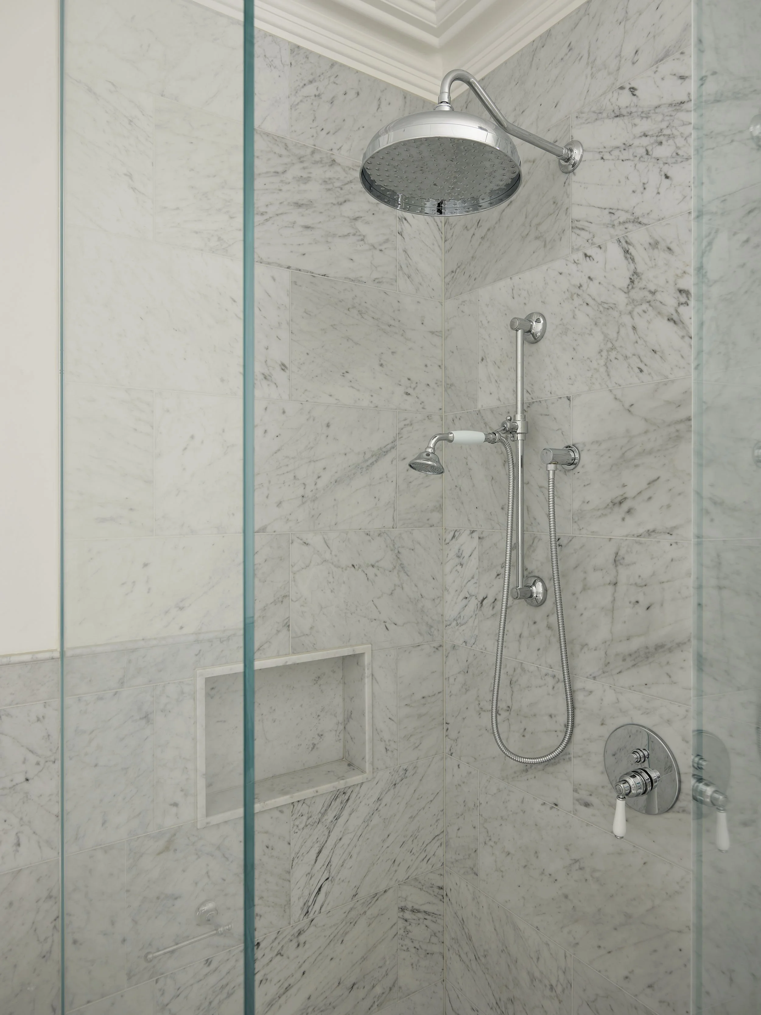 A marble tiled shower with a large rainfall showerhead, a handheld showerhead, and a built-in soap shelf.