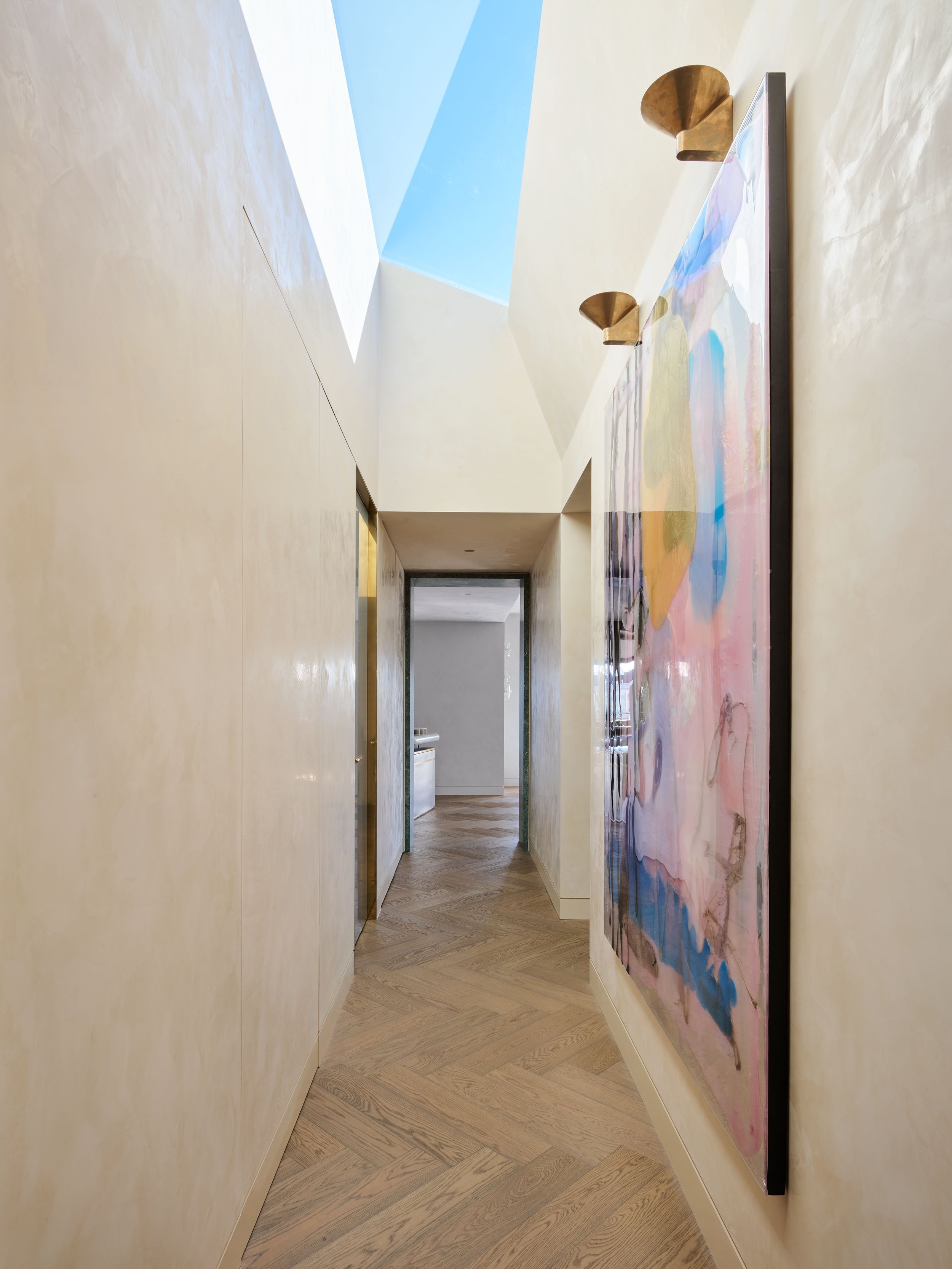A modern hallway with light-colored walls, wooden herringbone floor, artwork on the right wall, and skylights at the ceiling.