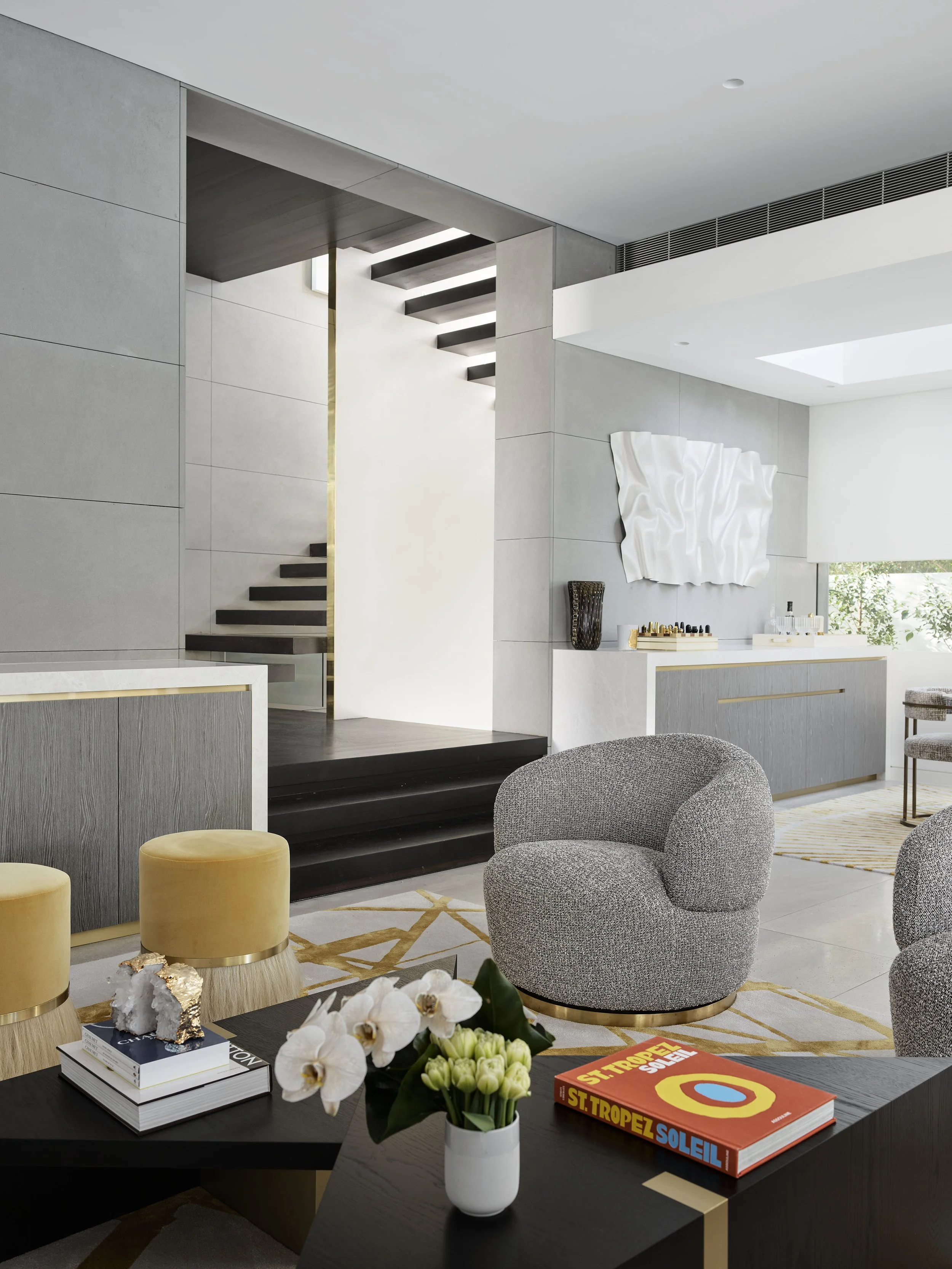 Modern living room with gray textured chairs, a black coffee table holding a white orchid, books, and decorative items, and furniture with gold accents. An abstract white wall art is visible on the wall, and the room features minimalist decor with ne
