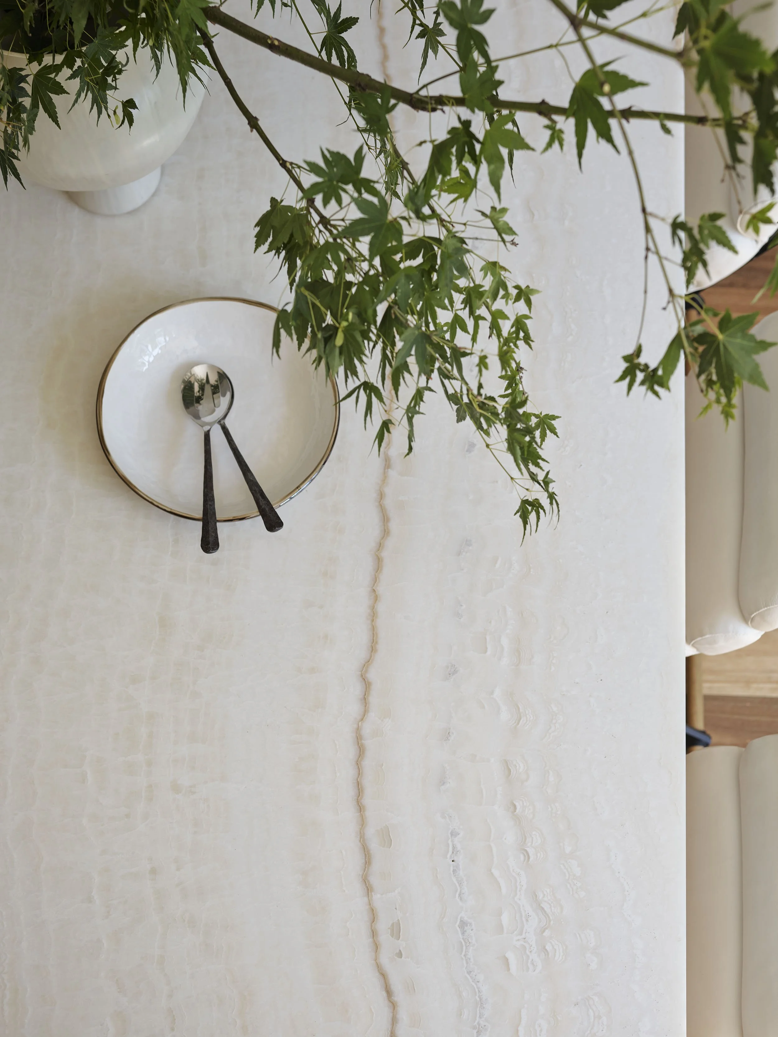 Top-down view of a dining table with a beige, marble-like surface, a white bowl containing two metal spoons, and a green leafy plant in a white ceramic vase.