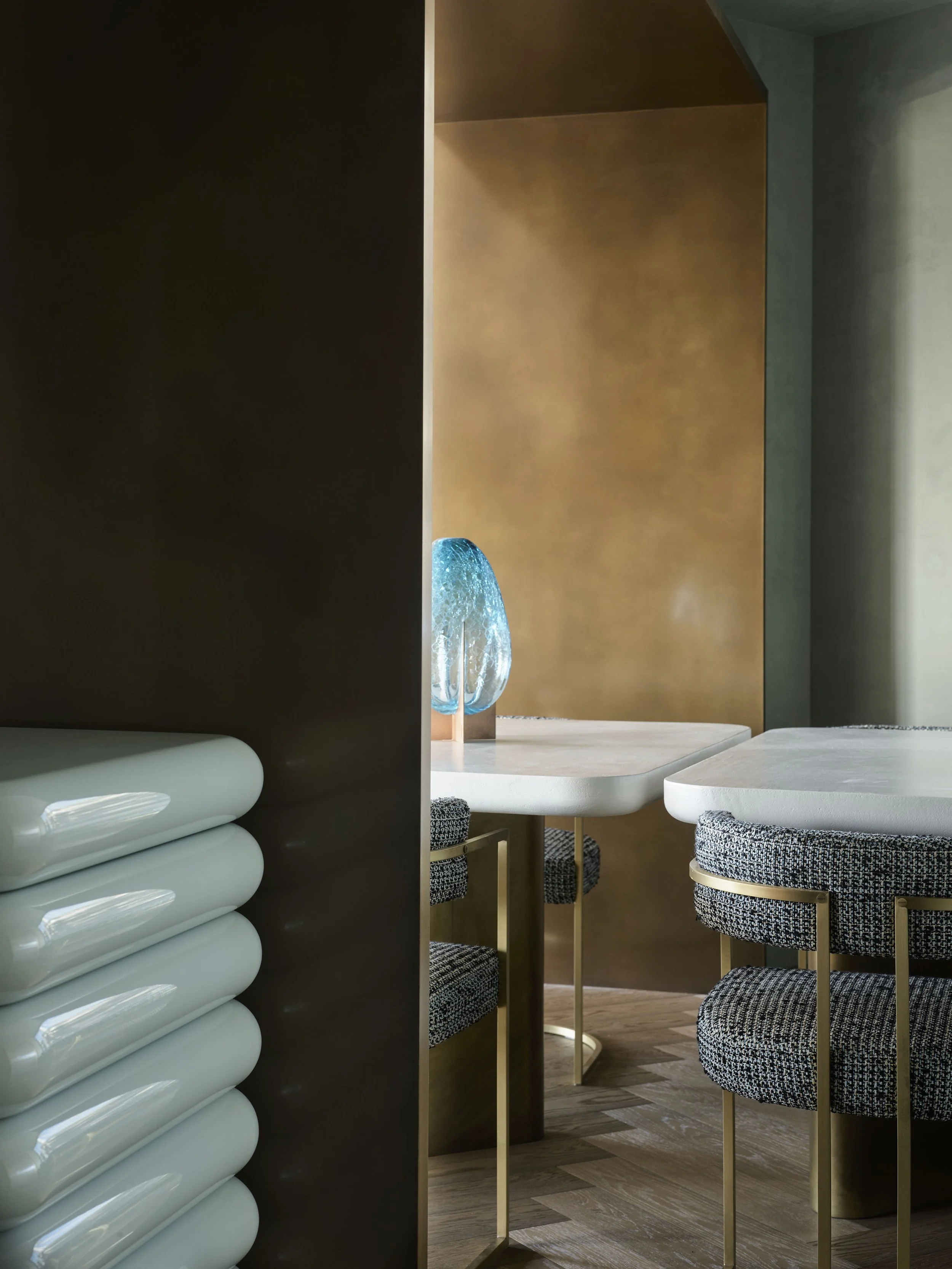 Interior view of a dining area with modern decor, including a shelf with white rolled towels, a glass sculpture on a table, and textured chairs with gold legs.