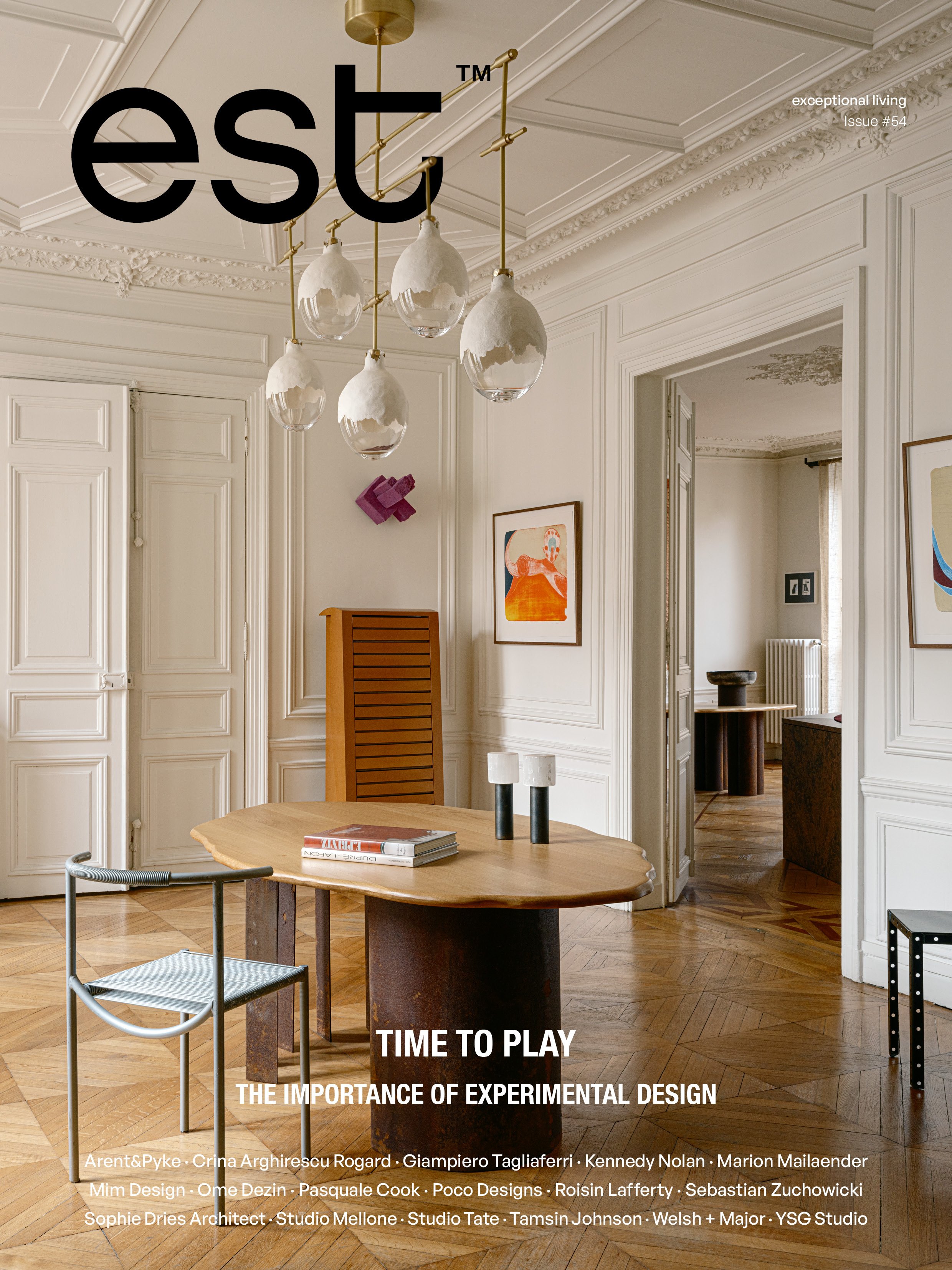 Cover of a magazine titled 'est' with the subtitle 'exceptional living, Issue #54'. The cover features a stylish room with ornate white walls, a wooden table, a modern chair, and a striking chandelier with glass and gold accents. There are several pieces of artwork on the walls.