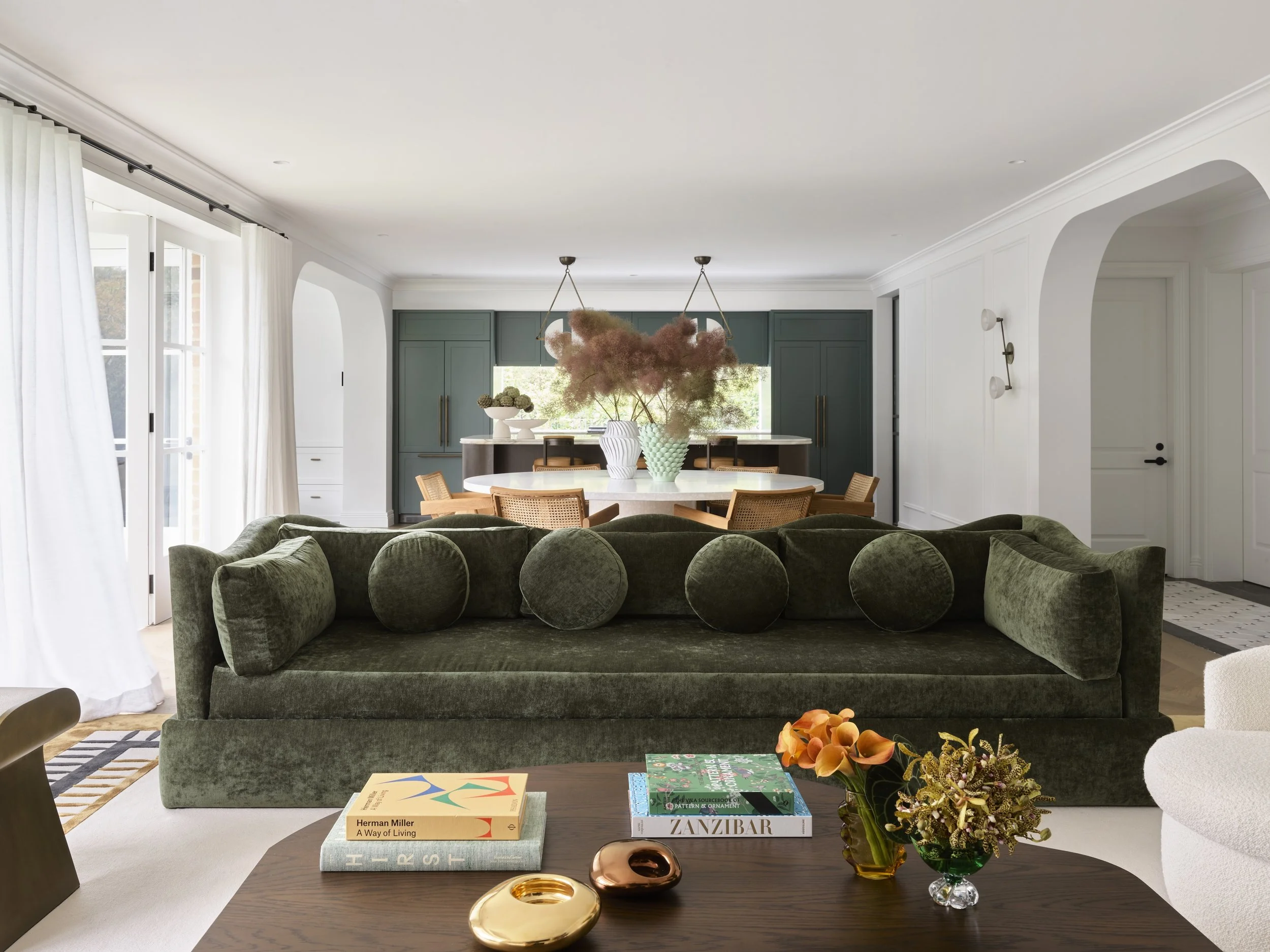 Living room with dark green velvet sofa, middle table with books, including 'Herman Miller A Way of Living' and 'Zanzibar', floral arrangement, white armchair, open view to dining area with a round table, green cabinetry, and pink floral chandelier.