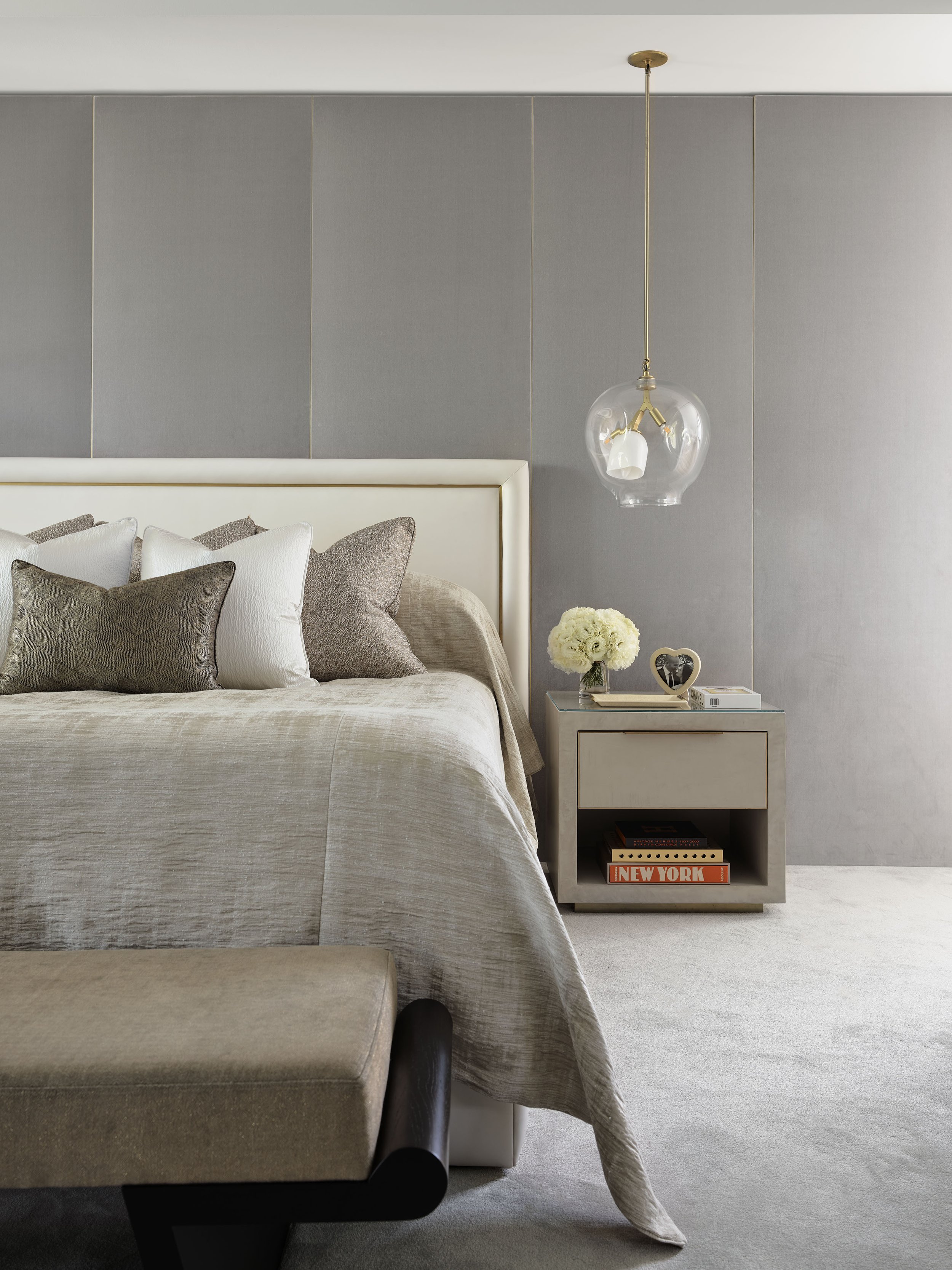 A bedroom with a beige bed, decorative pillows, a nightstand with a flower bouquet, a picture frame, and books, a modern glass pendant light, and a gray-paneled wall.