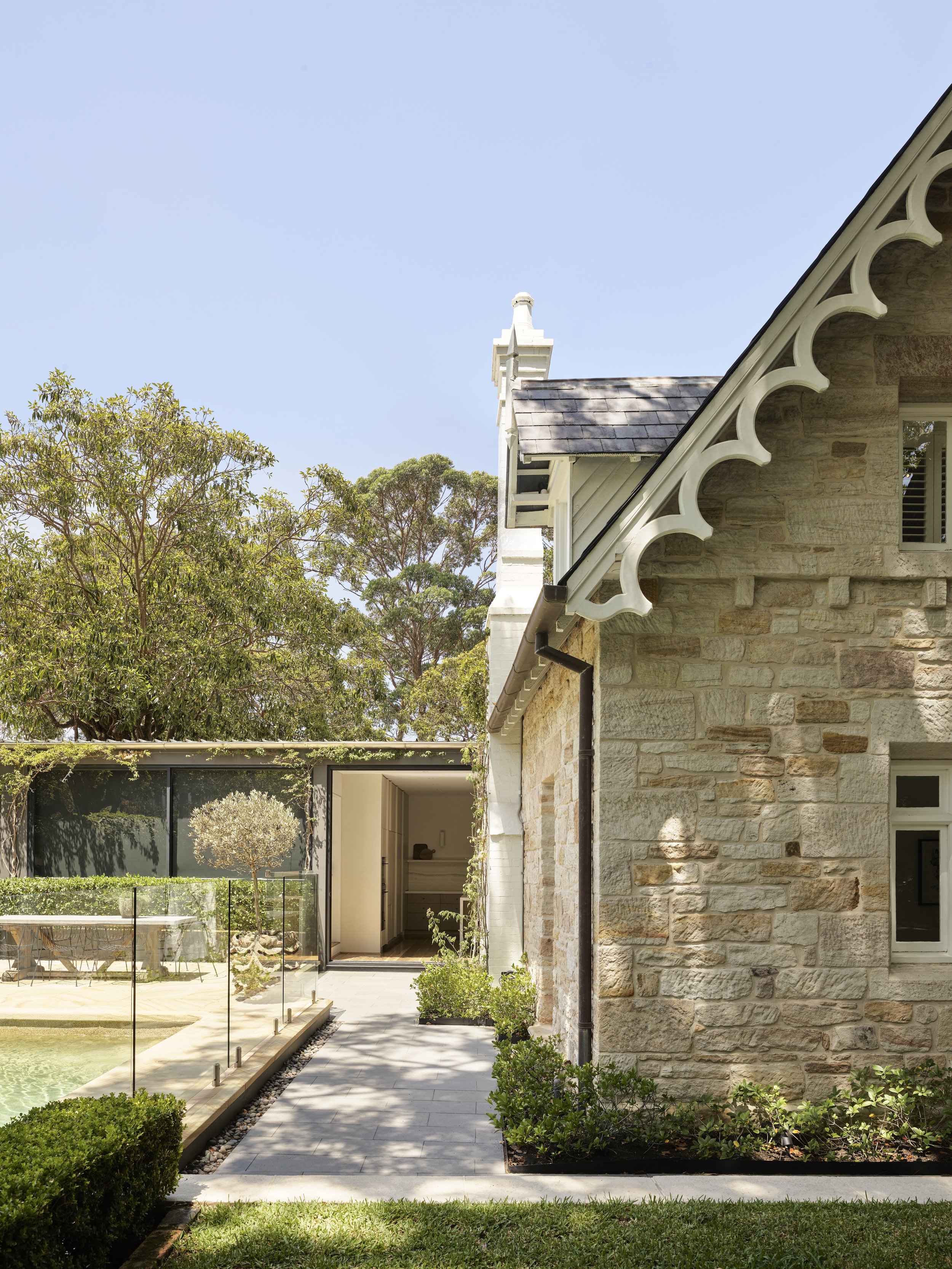 Side view of a stone house with decorative white trim on the roof edge, adjacent to a landscaped yard with a pathway, bushes, and a small pool with a glass fence.