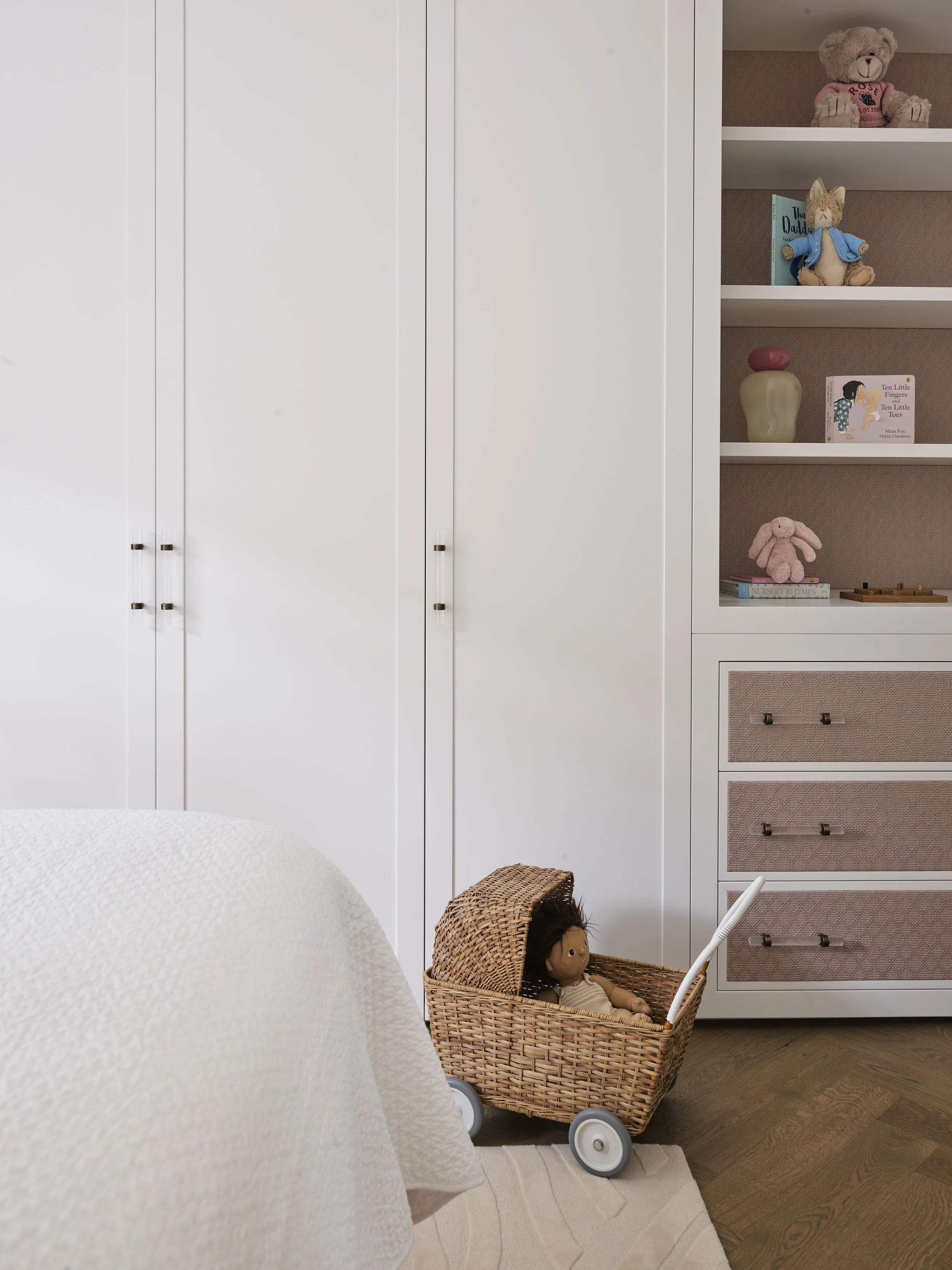 A corner of a bedroom with a white wardrobe, a bookshelf with plush toys and books, and a wicker doll stroller with a doll inside.