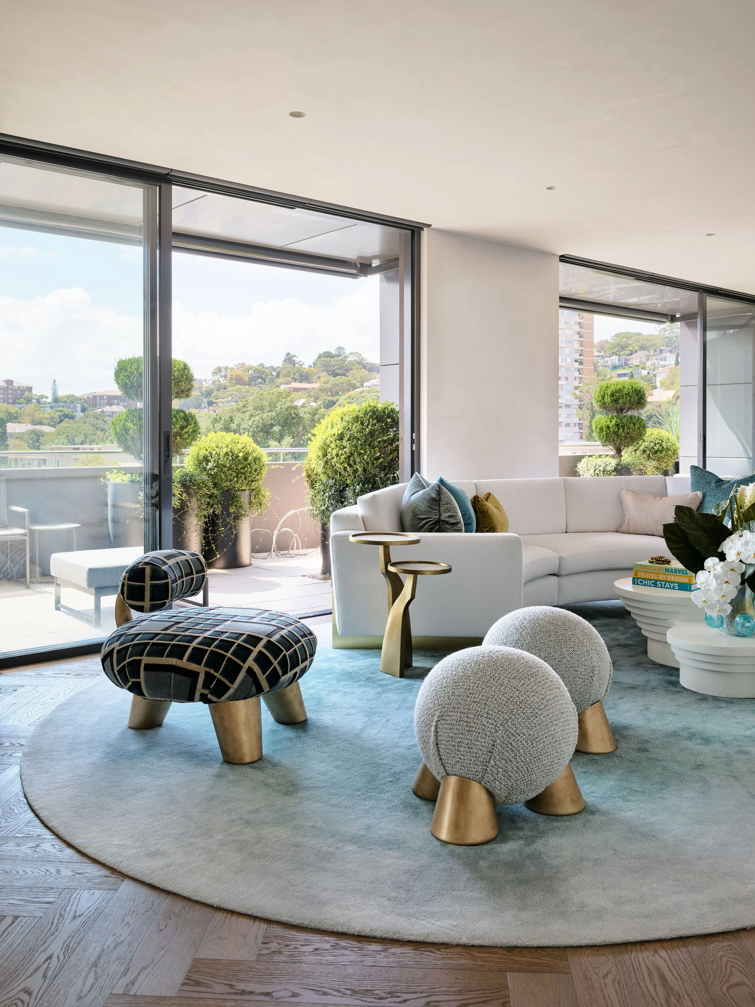 Modern living room with a white sofa, decorative pillows, and a round coffee table with books. Front view of a sliding glass door leading to a balcony with potted plants and outdoor seating. Colorful stool and spherical ottomans on a circular rug, wi