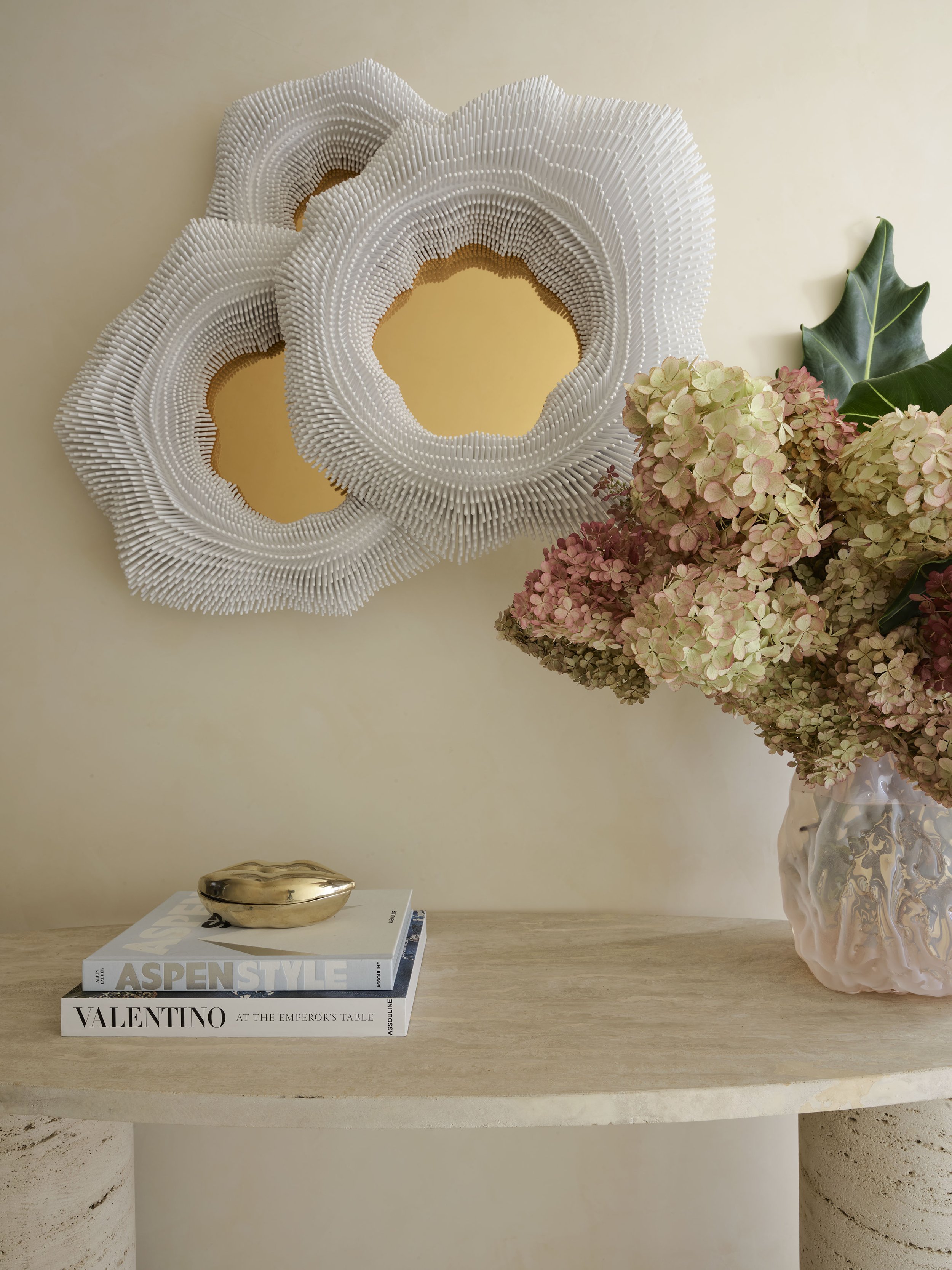 Decorative wall art with layered white circular shapes on a yellow background, beside a bouquet of pink hydrangeas in a textured ceramic vase on a wooden table, with a gold-colored dish and books titled 'Aspen Style' and 'Valentino'.