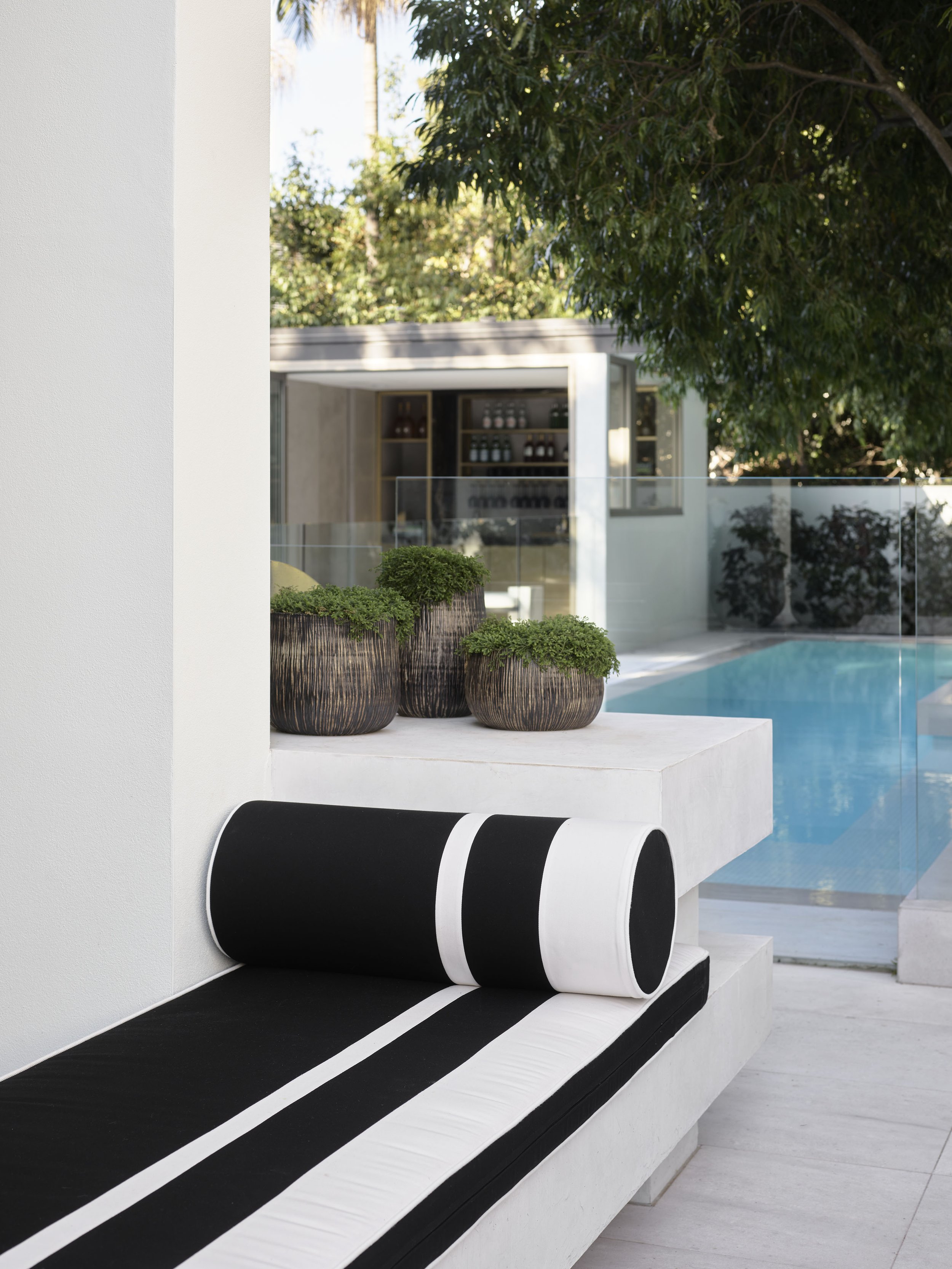 Modern outdoor patio with black and white striped lounge chair, potted plants, swimming pool, and glass pool fence, surrounded by trees.